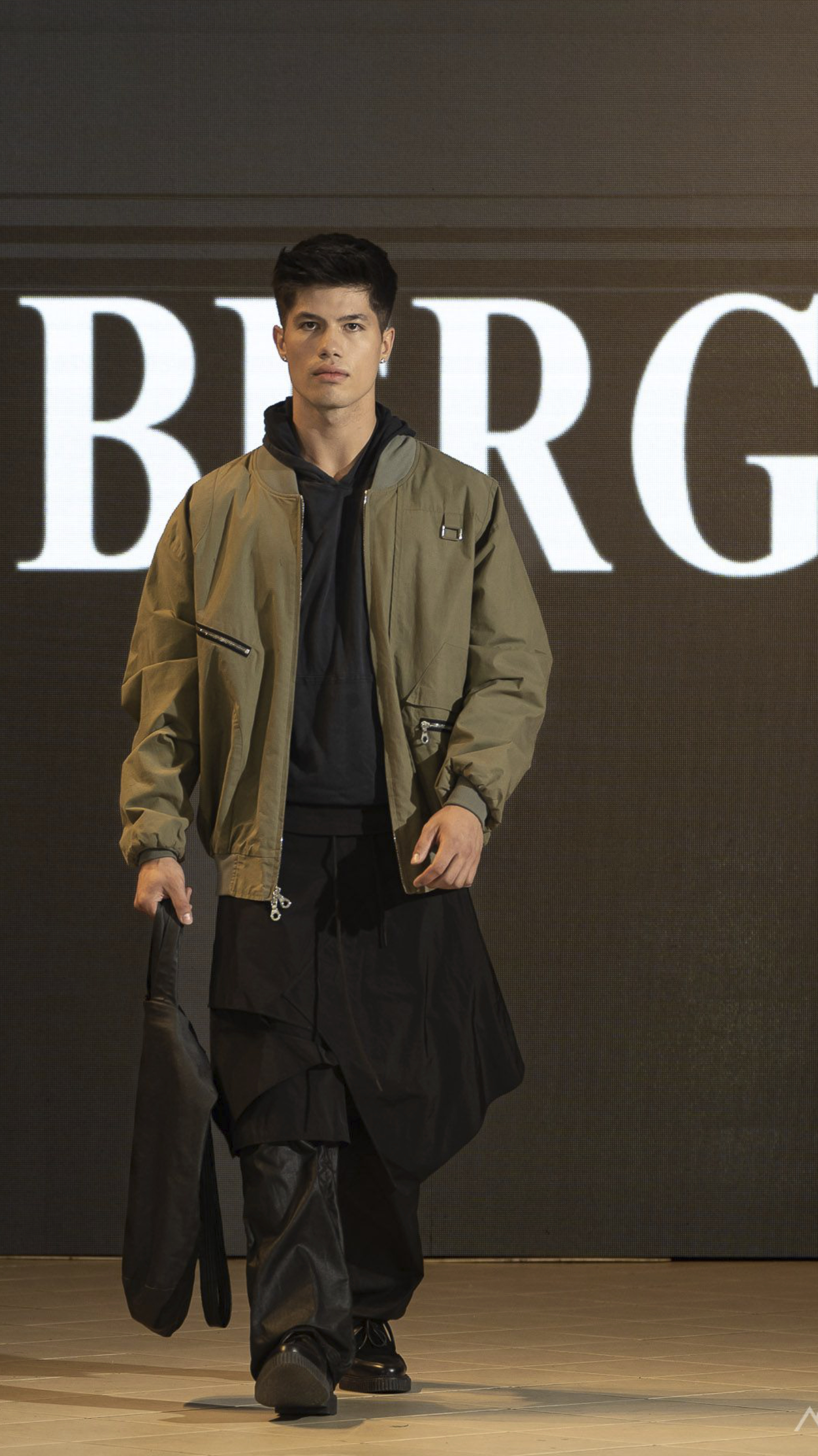 Male model walking down runway wearing an olive green bomber jacket, black hoodie, layered black skirt over black pants, holding a black bag.