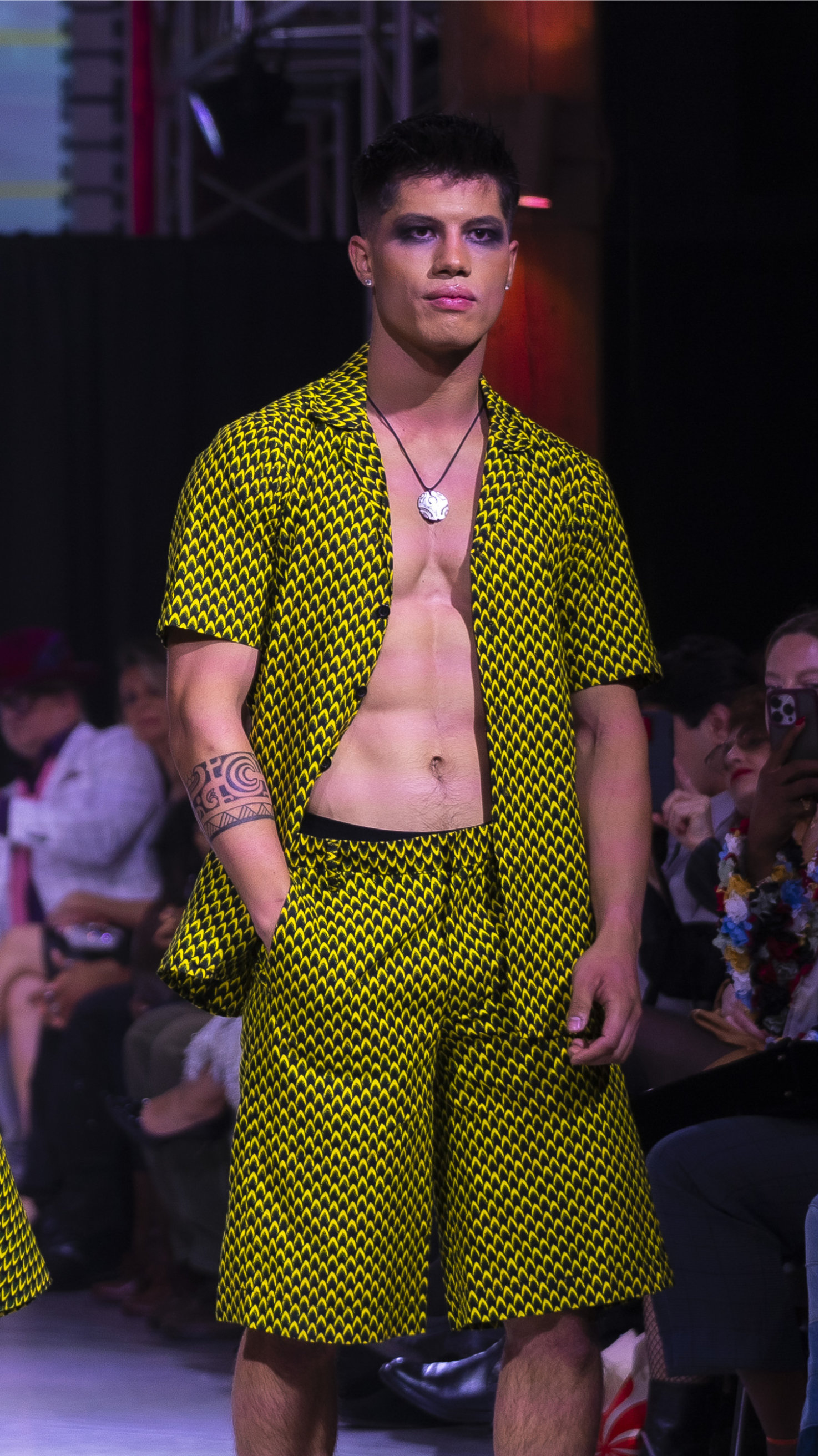 Male model with dark makeup wearing a yellow and black patterned short-sleeve shirt and matching shorts on a fashion runway.