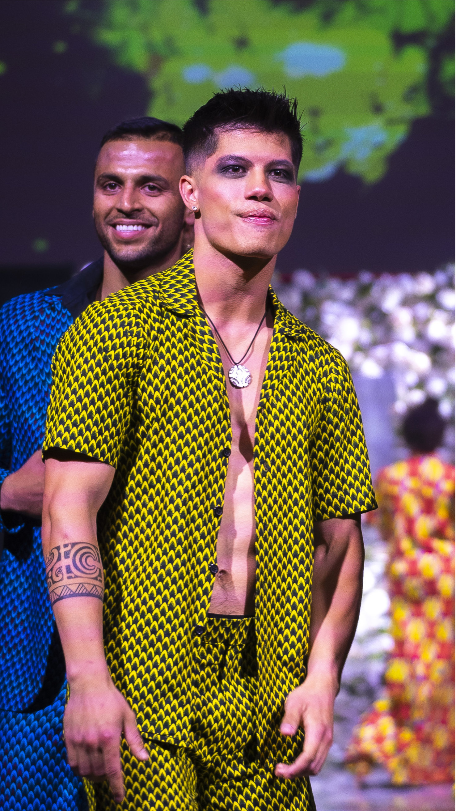 Two male models on a runway, one wearing a yellow patterned open shirt and matching pants, the other in a blue patterned outfit.