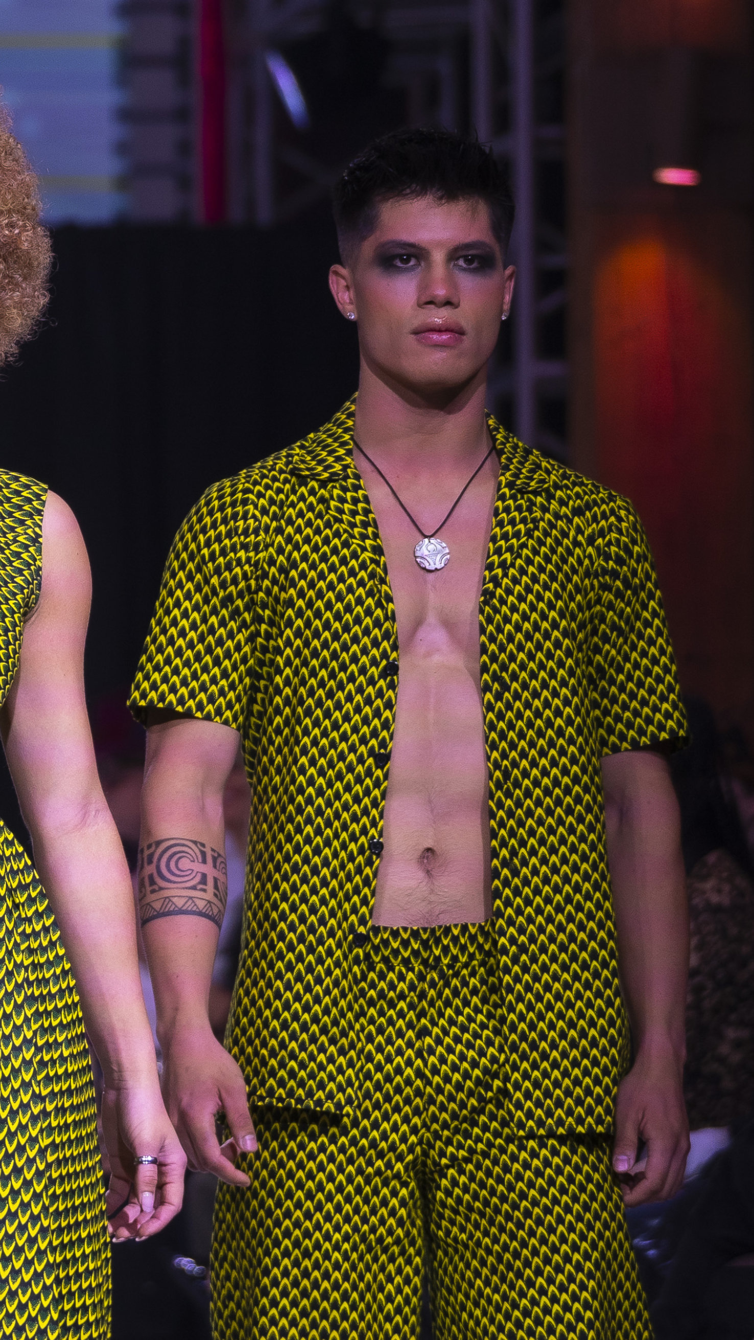 Male model with dark eye makeup wearing a yellow and black patterned open shirt and matching shorts with a tattoo on his left forearm.