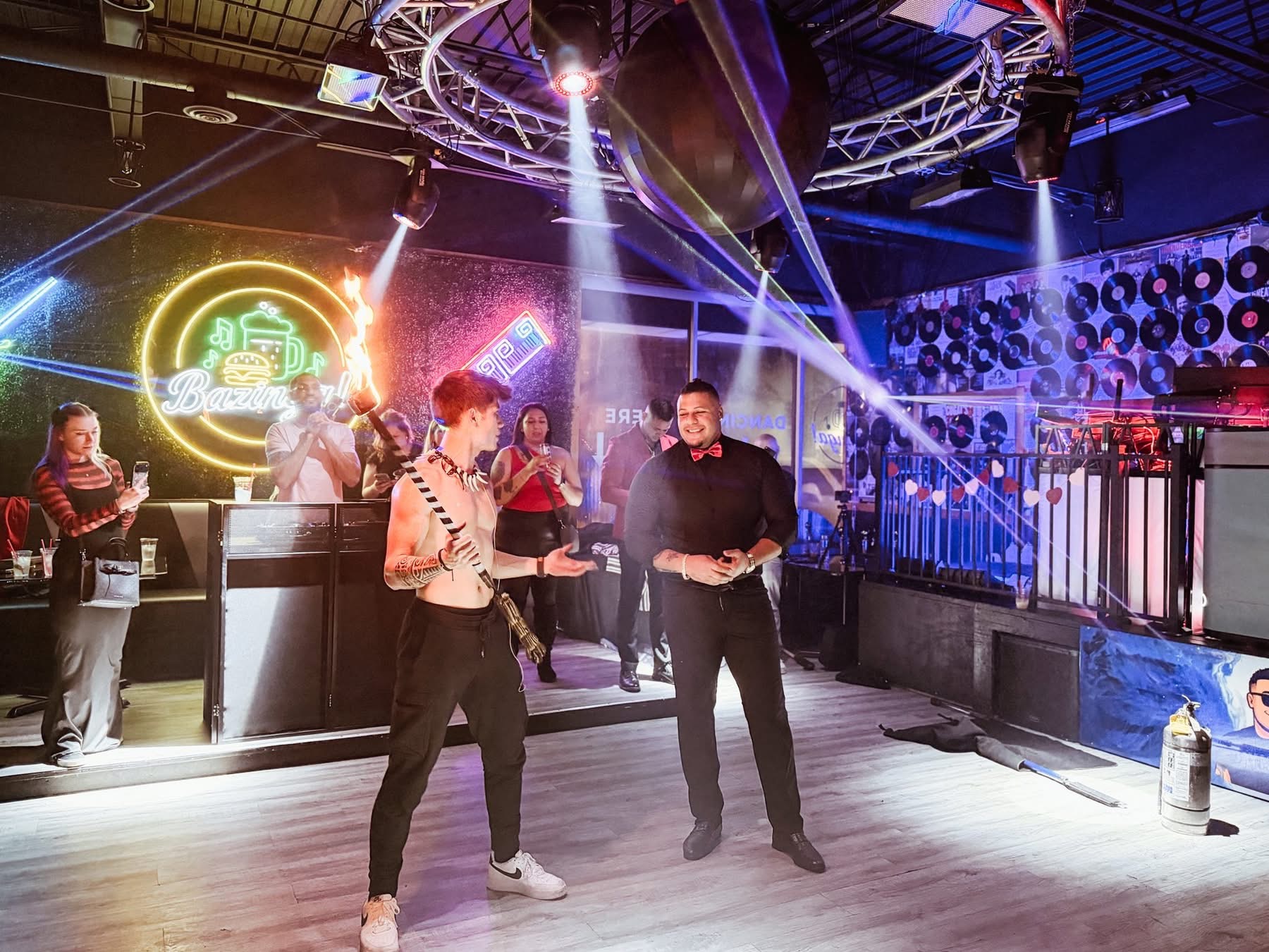 Two men standing in a room with neon signs and vinyl records on the wall, one holding a flaming torch, surrounded by people watching and taking photos under bright stage lights.