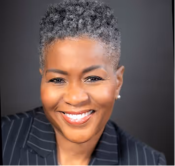 Smiling woman with short gray hair wearing a black pinstripe blazer against a dark background.