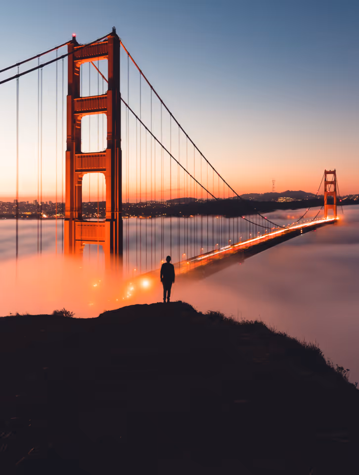 Silhouette of a person standing on a hill overlooking the illuminated Golden Gate Bridge above fog at sunset.