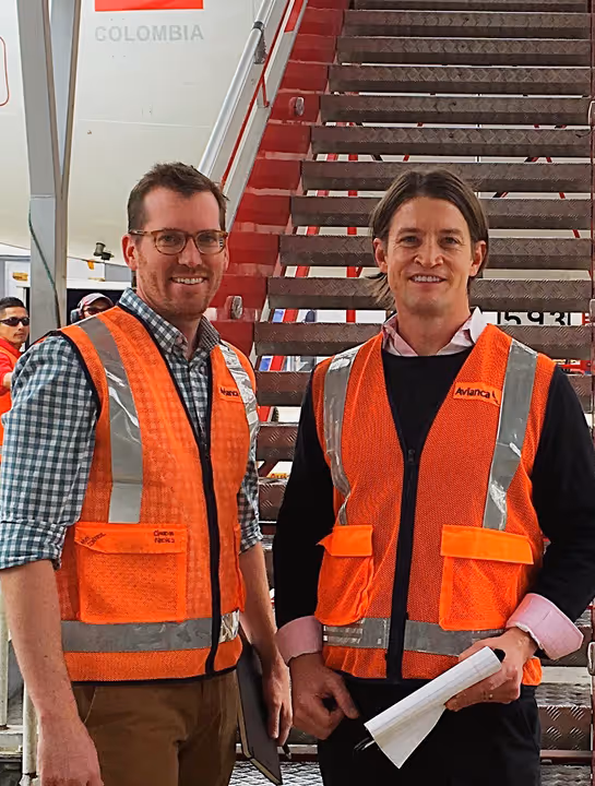 Two men wearing orange safety vests standing in front of an airplane boarding staircase labeled Colombia.