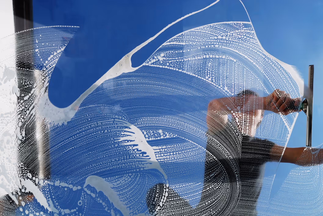 Man cleaning a window with a squeegee, viewed through soapy water and suds against a blue sky.