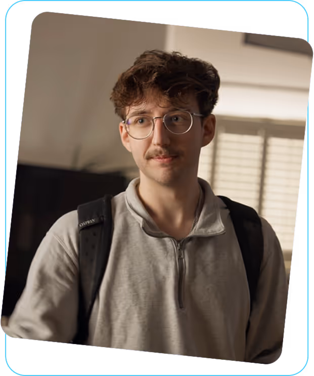 Young man with glasses and mustache wearing a light gray zip-up sweater and a black backpack indoors.