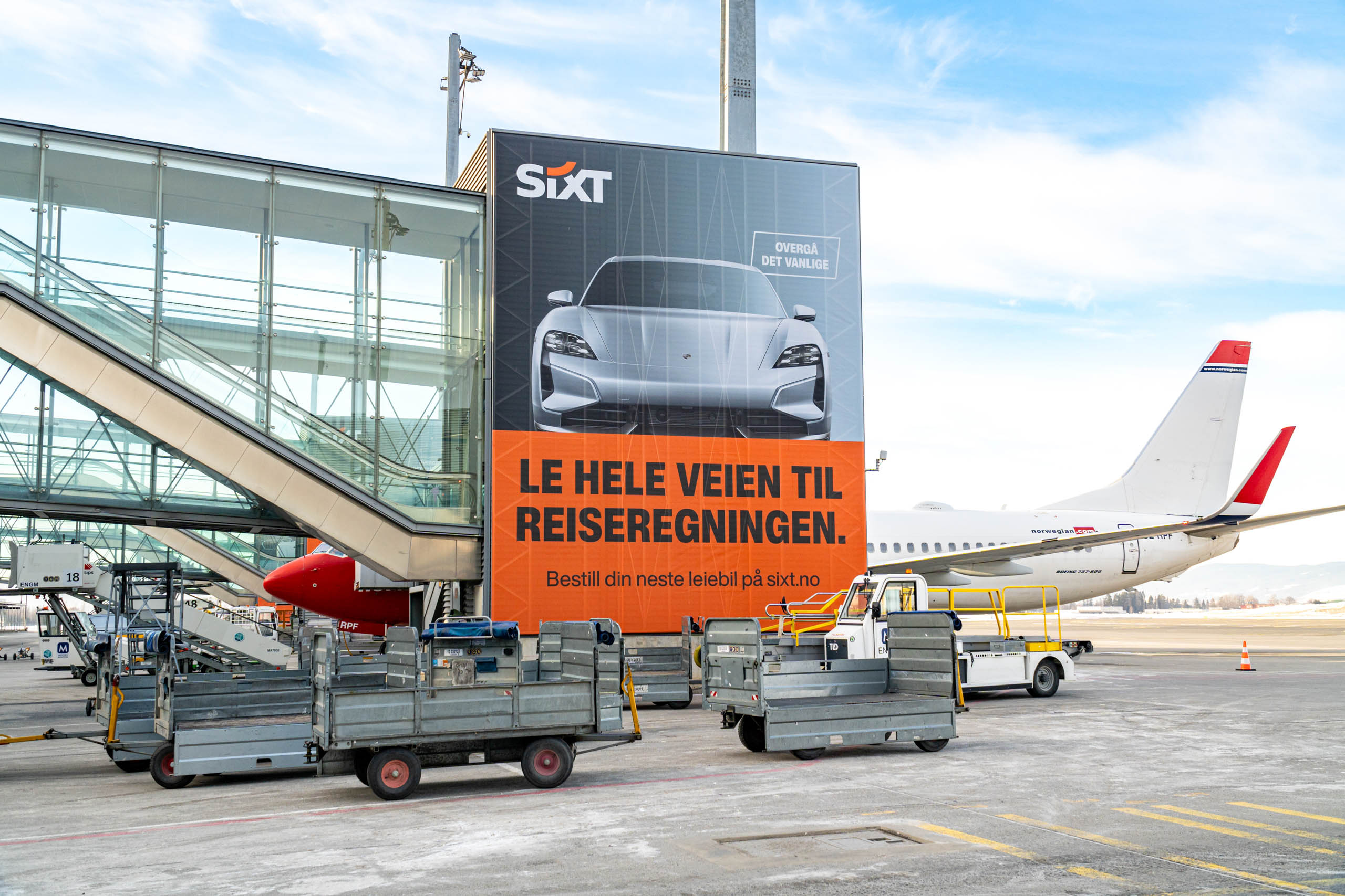 Reklameskilt for leiebil foran en Norwegian-flygate med bagasjetraller og flybro.