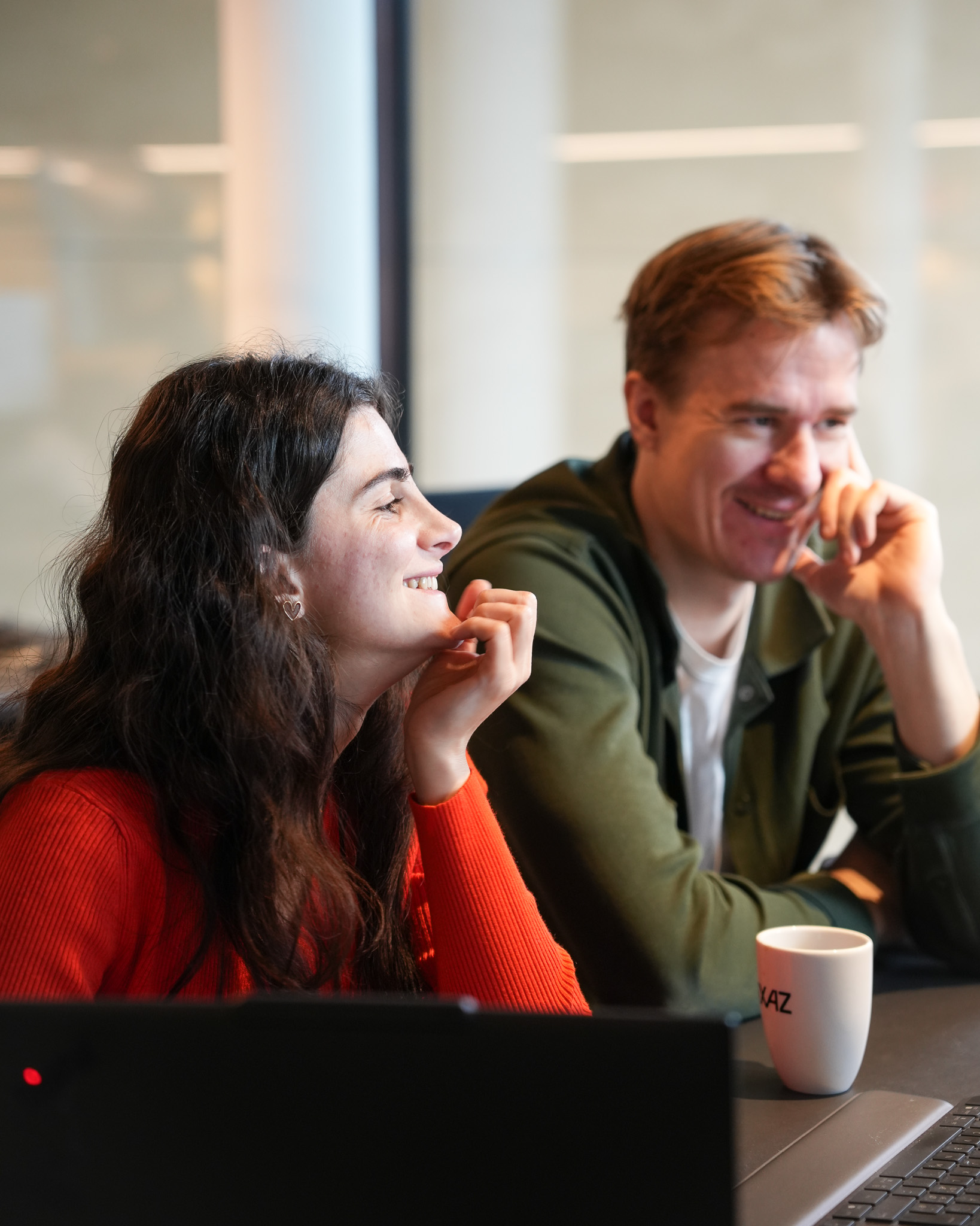 To personer smilende og ser mot en dataskjerm i et kontormiljø.