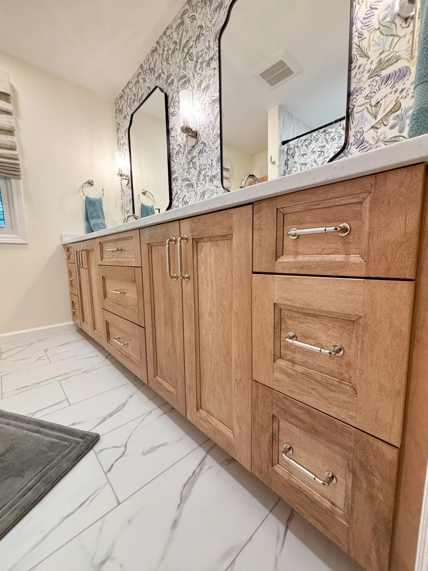 Luxurious transitional bathroom with wood cabinetry, double sinks, marble tile flooring, and floral wallpaper.
