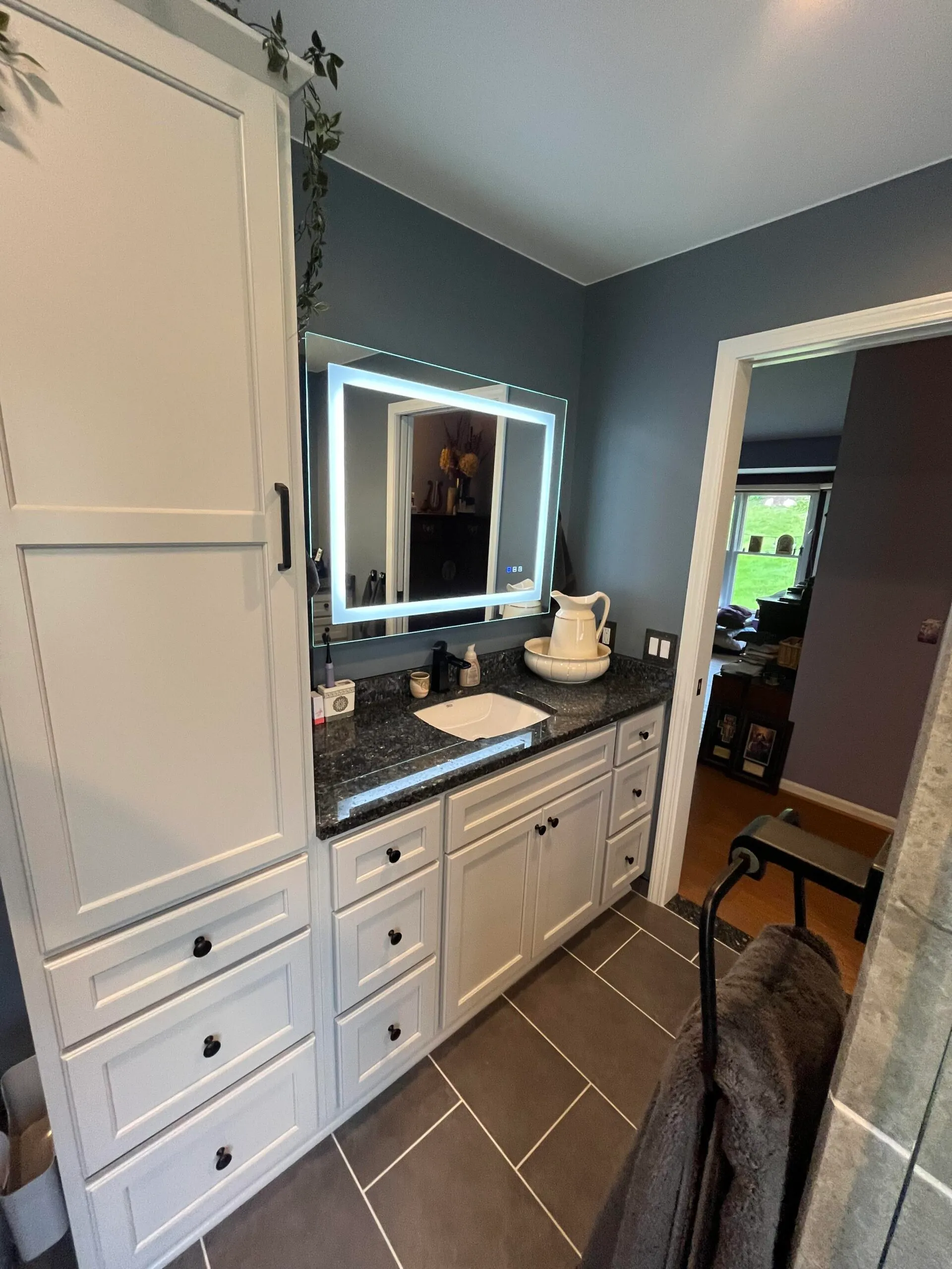 A modern bathroom featuring a sleek white vanity with a square LED mirror, gray walls, and dark tile flooring.