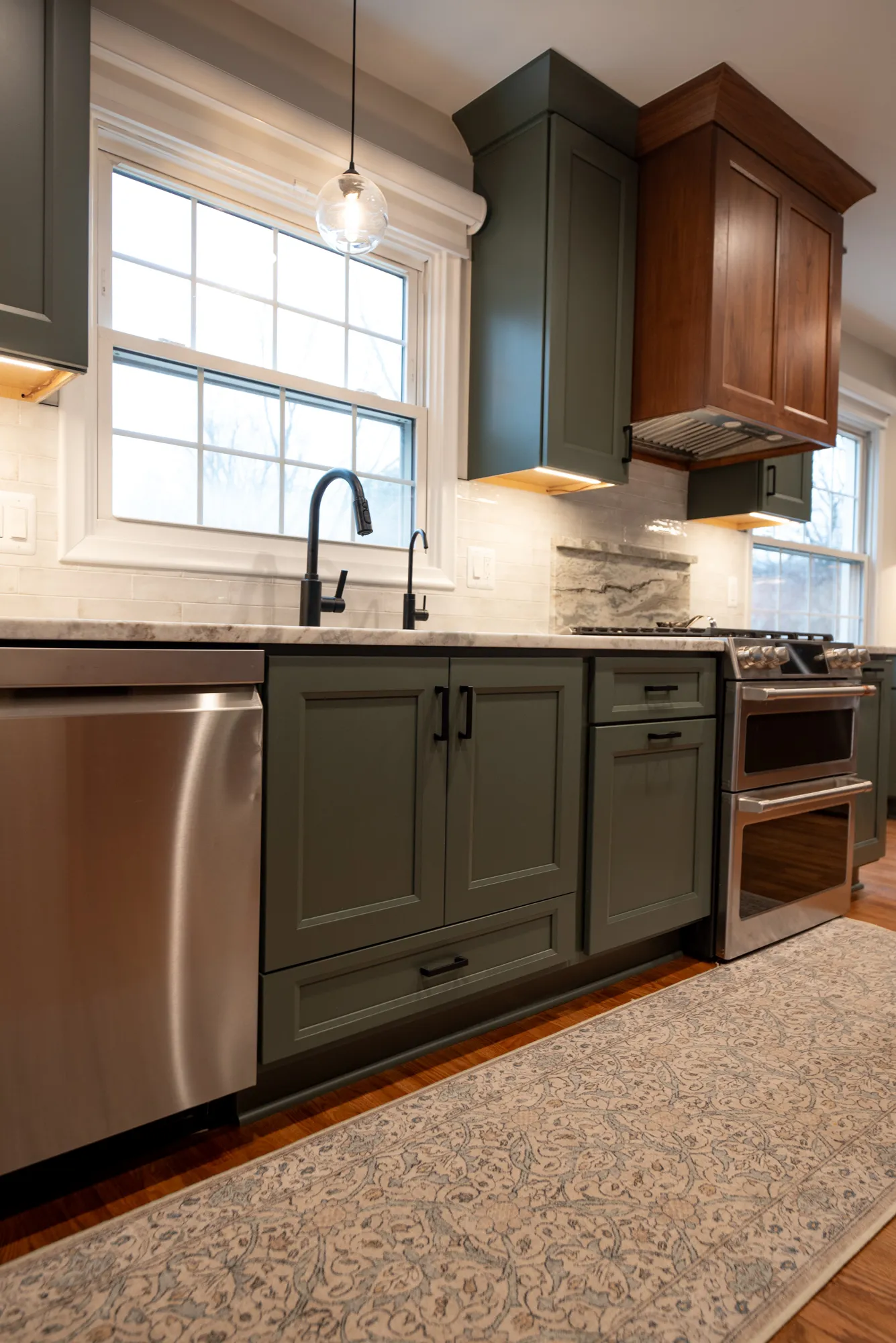 Modern kitchen with dark green cabinets, marble countertops, and stainless steel appliances, featuring a decorative rug.