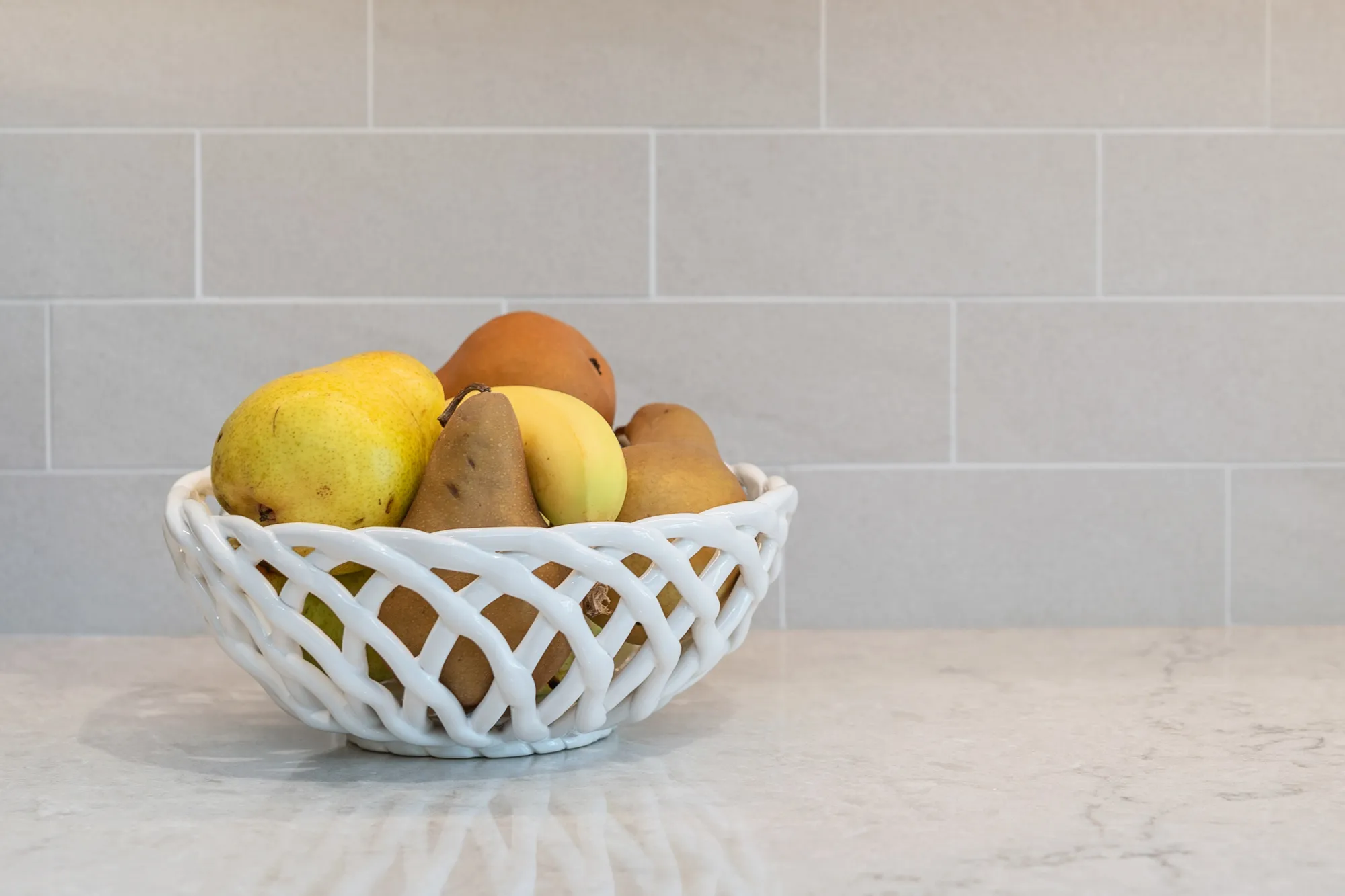 Fruit bowl filled with pears and bananas on a marble countertop in a kitchen with gray tile backsplash
