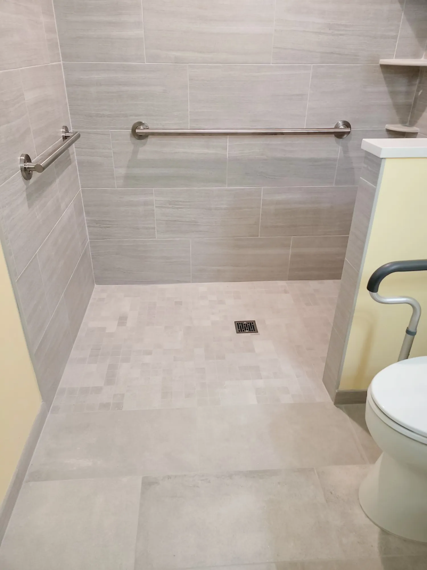 Modern bathroom shower with gray tile walls, textured flooring, and grab bars for safety