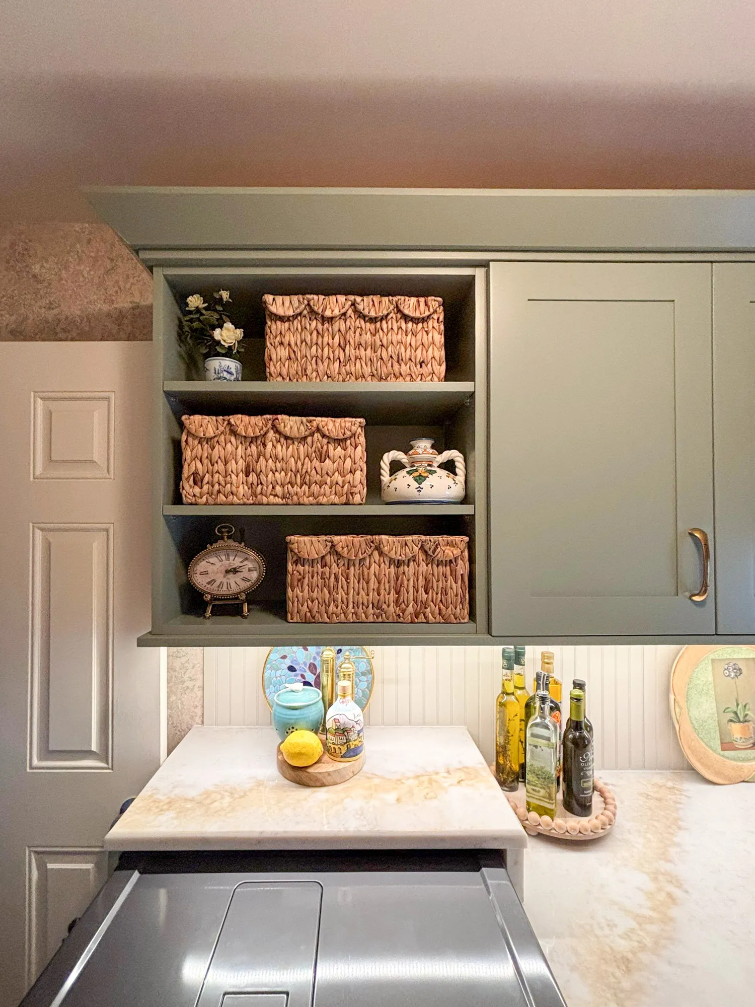 Charming kitchen with green cabinets, vintage decor, and a marble countertop showcasing olive oil bottles and decorative plates.