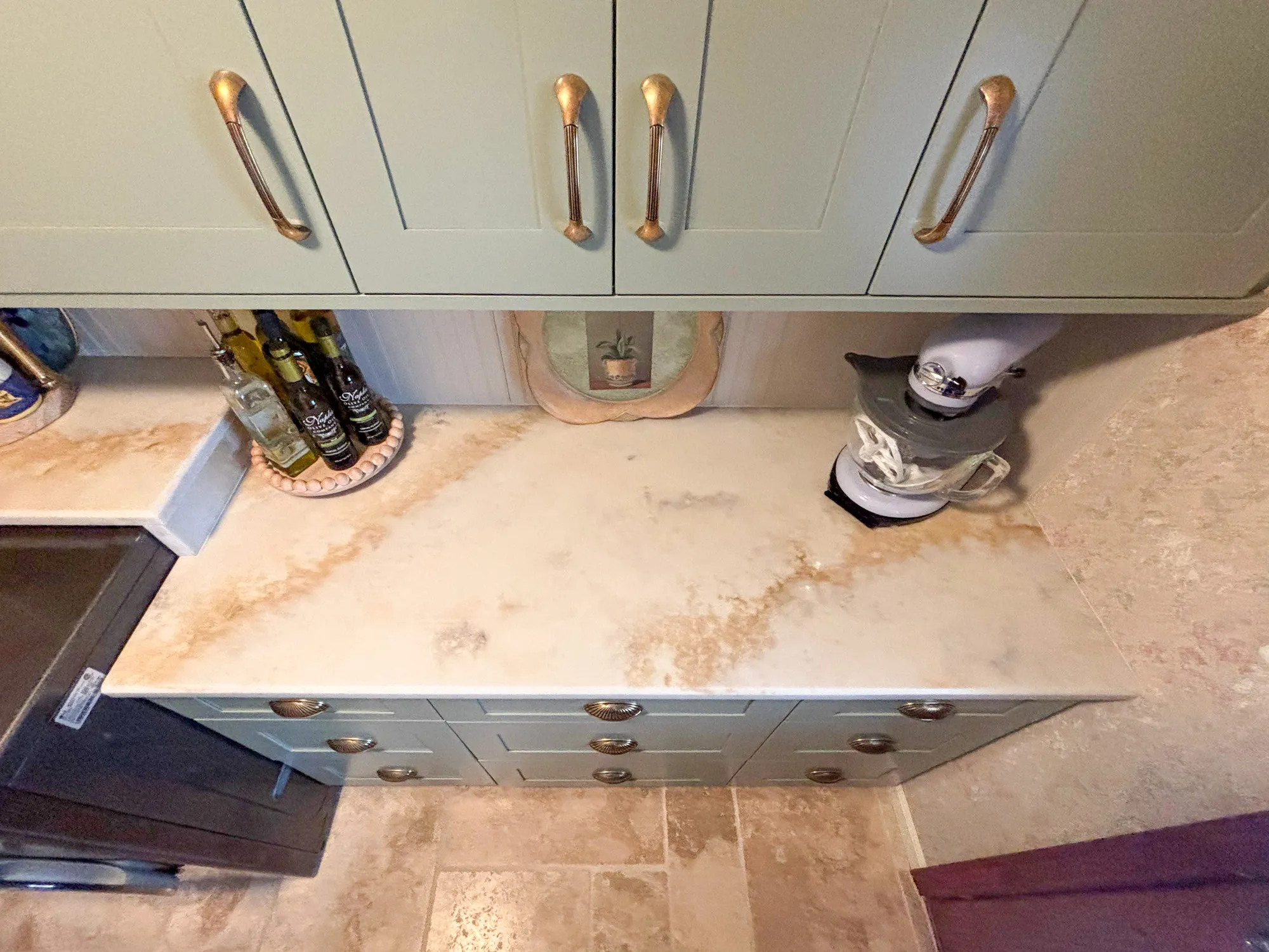 Contemporary kitchen with pale green cabinetry and marble countertops features a mixer and decorative oil bottles.