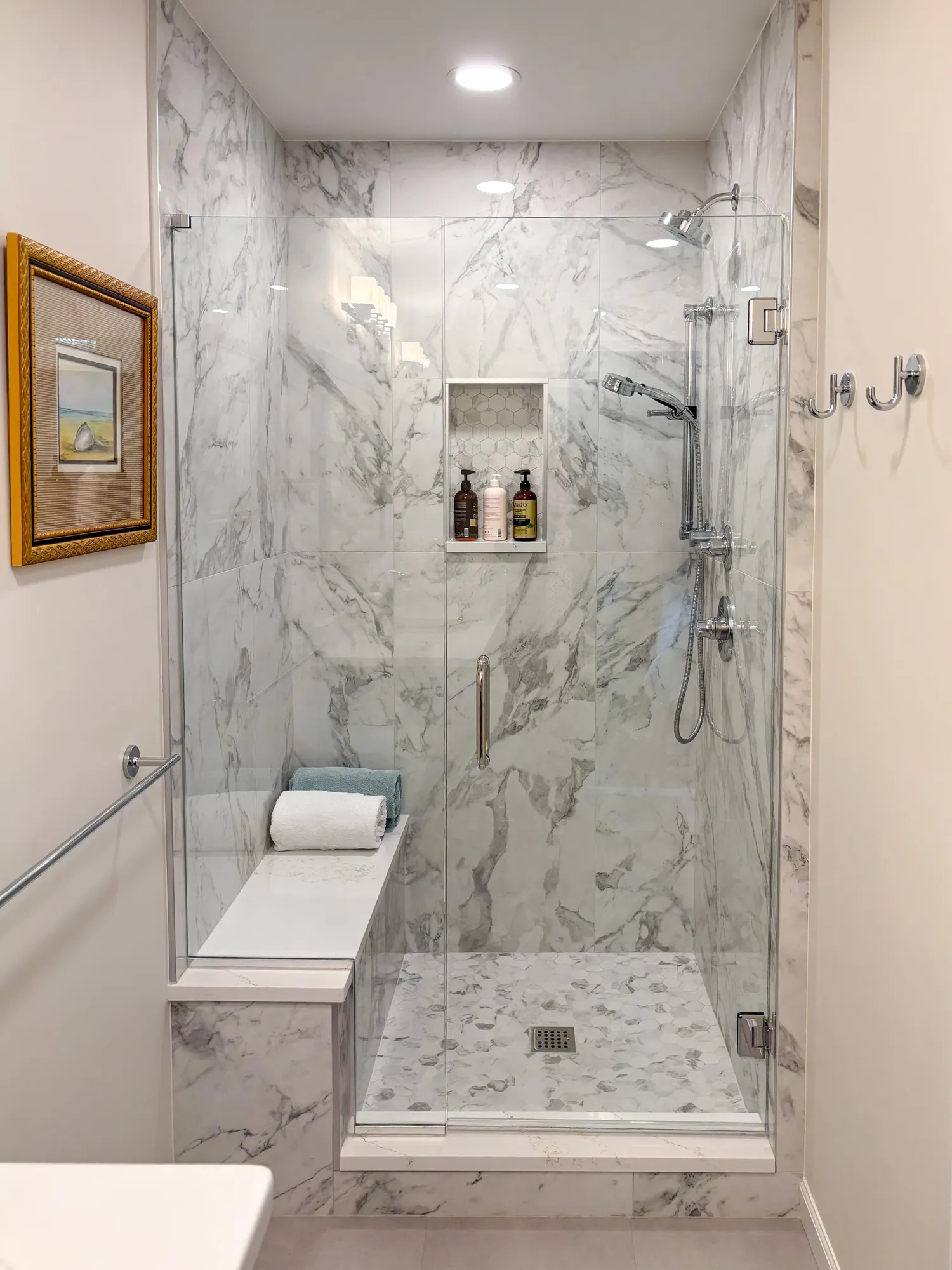 Modern bathroom shower with marble walls and glass enclosure, featuring a shelf and luxury toiletries