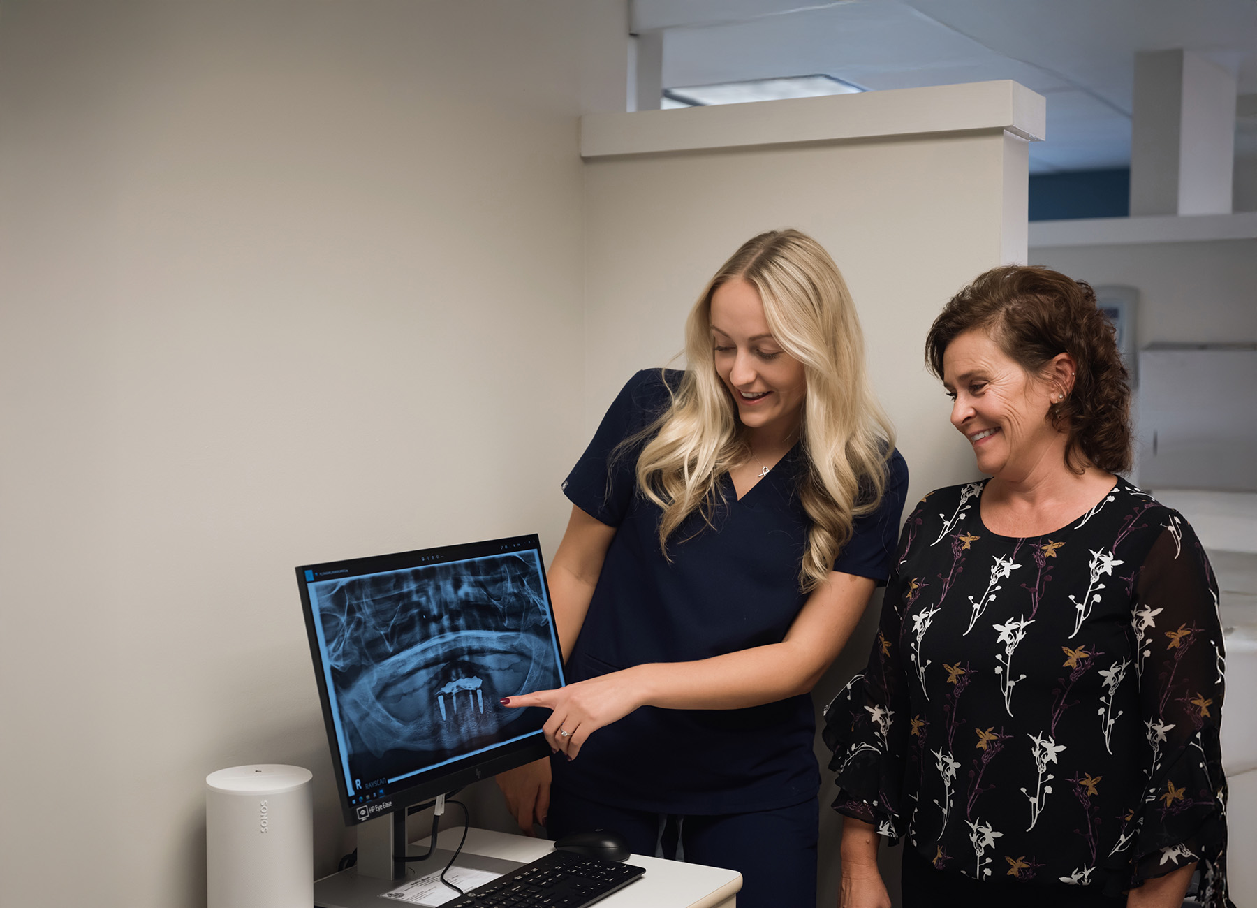 Dental professional in navy scrubs explaining dental X-ray on computer screen to a smiling patient in a black floral blouse.