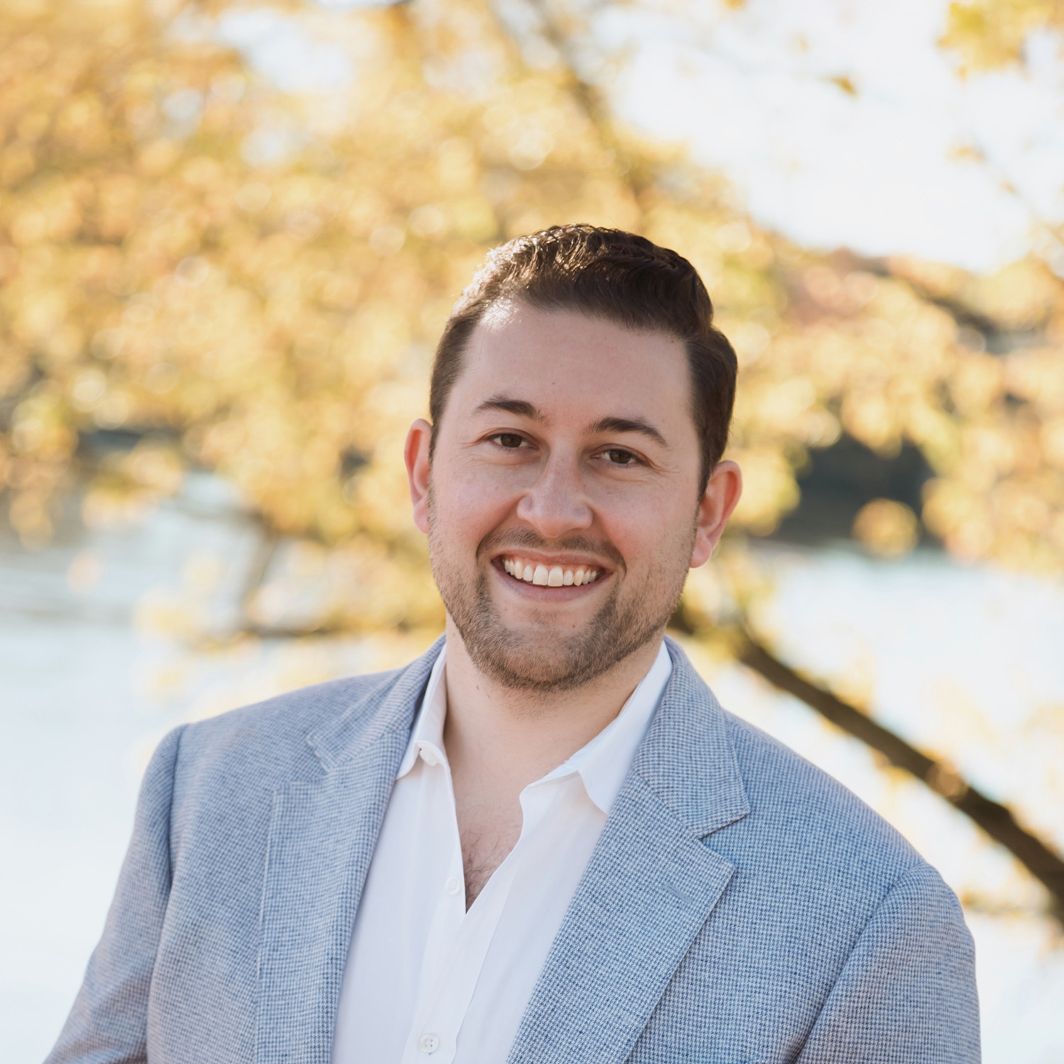 Smiling man with short dark hair and beard wearing a light gray blazer and white shirt outdoors with blurred yellow foliage background.
