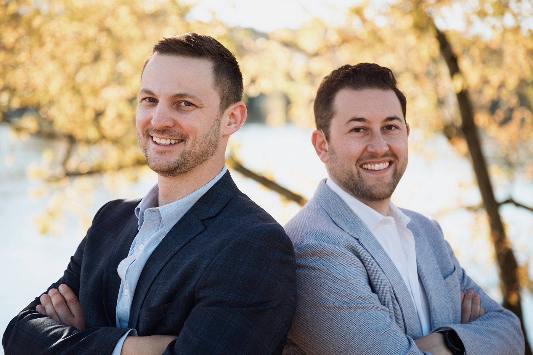 Two men smiling with arms crossed, standing outdoors with trees and water in the background.