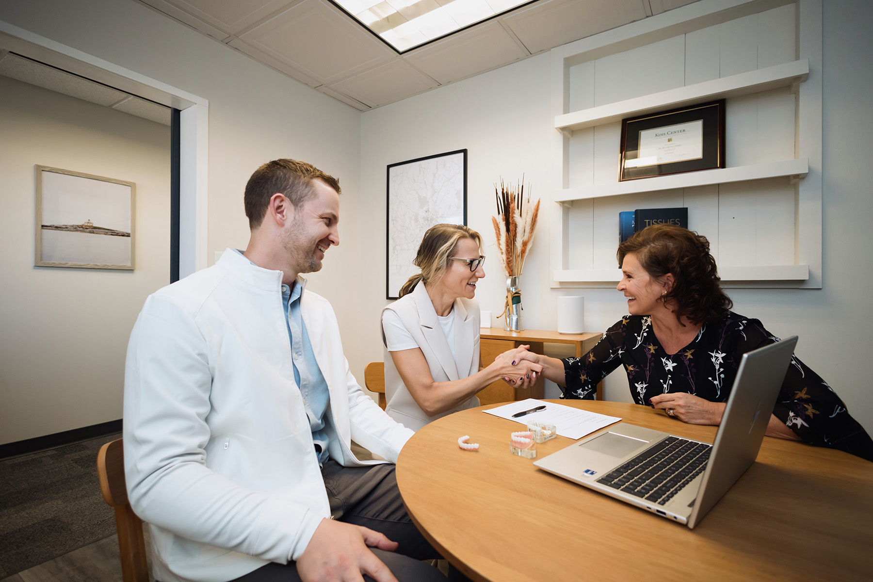 Two women shaking hands across a table with dental models and a laptop, while a man in a white jacket smiles beside them in an office.