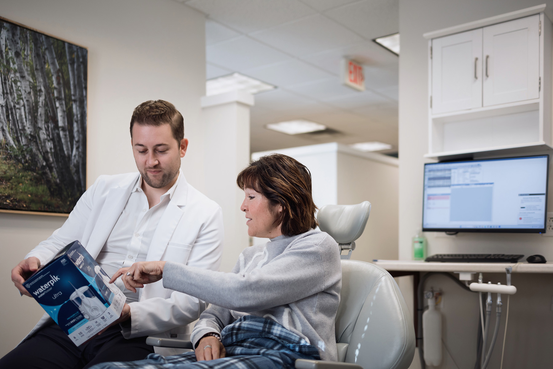 Dentist showing a Waterpik Ultra oral irrigator box to a female patient seated in a dental chair.
