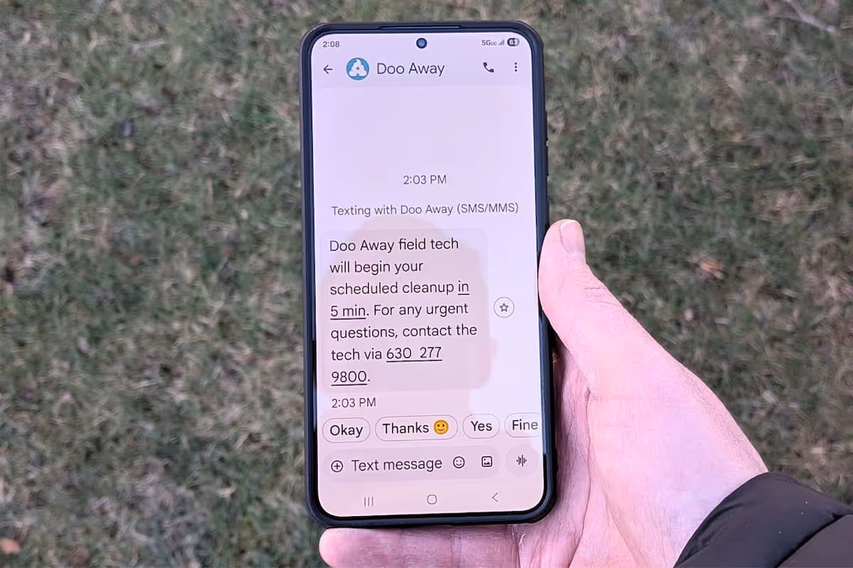 Hand holding a smartphone displaying a text message from Doo Away about scheduled cleanup starting in 5 minutes with a contact phone number.