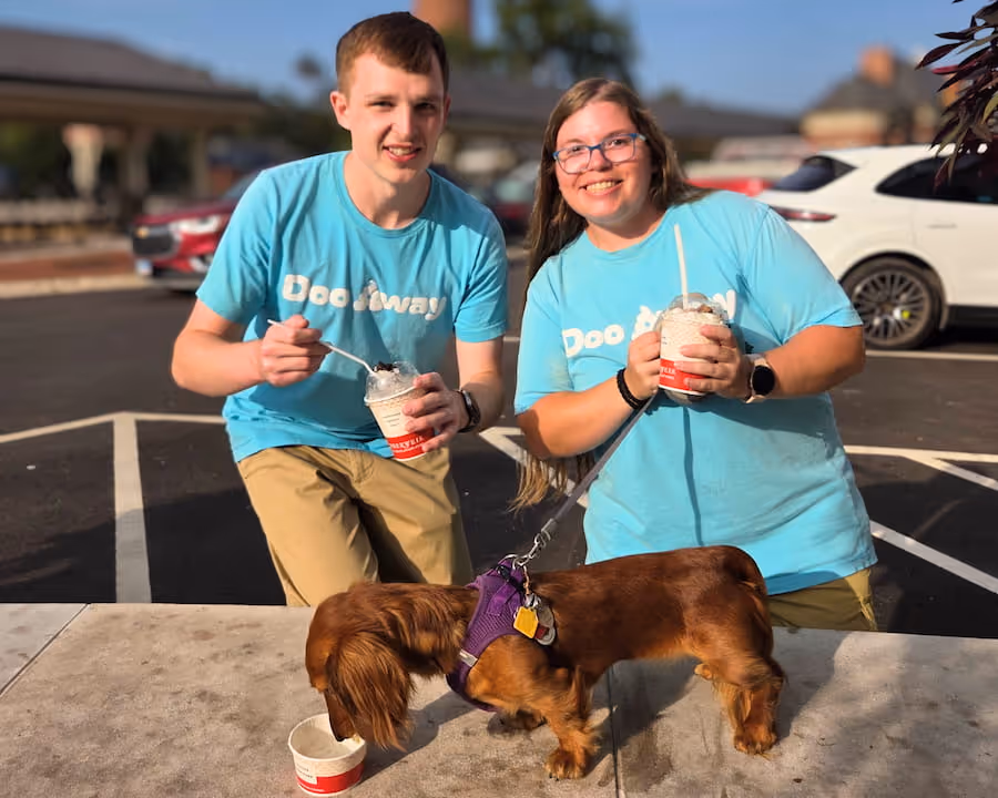 Doo Away team members in matching blue uniform smiling and eating ice cream with brown Dachshund on cement wall in front eating ice cream