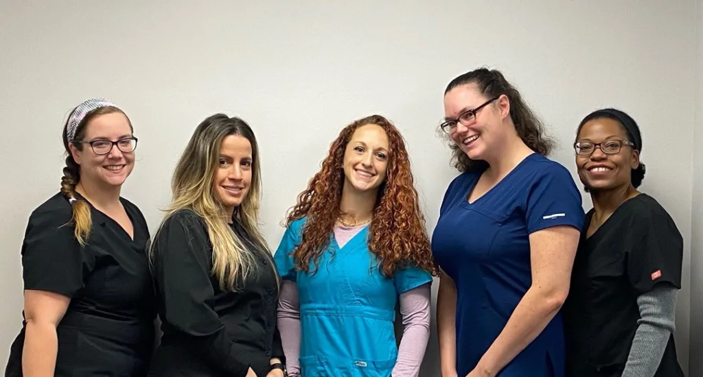 Group of five smiling female healthcare workers wearing scrubs standing against a plain wall.