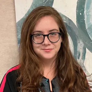 Young woman with long brown hair and black glasses smiling, wearing a black and red top.
