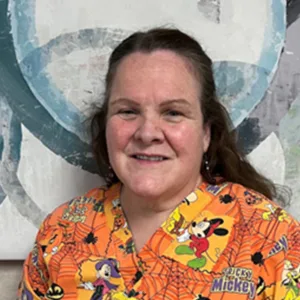 Smiling woman with long brown hair wearing an orange Mickey Mouse-themed scrub top.