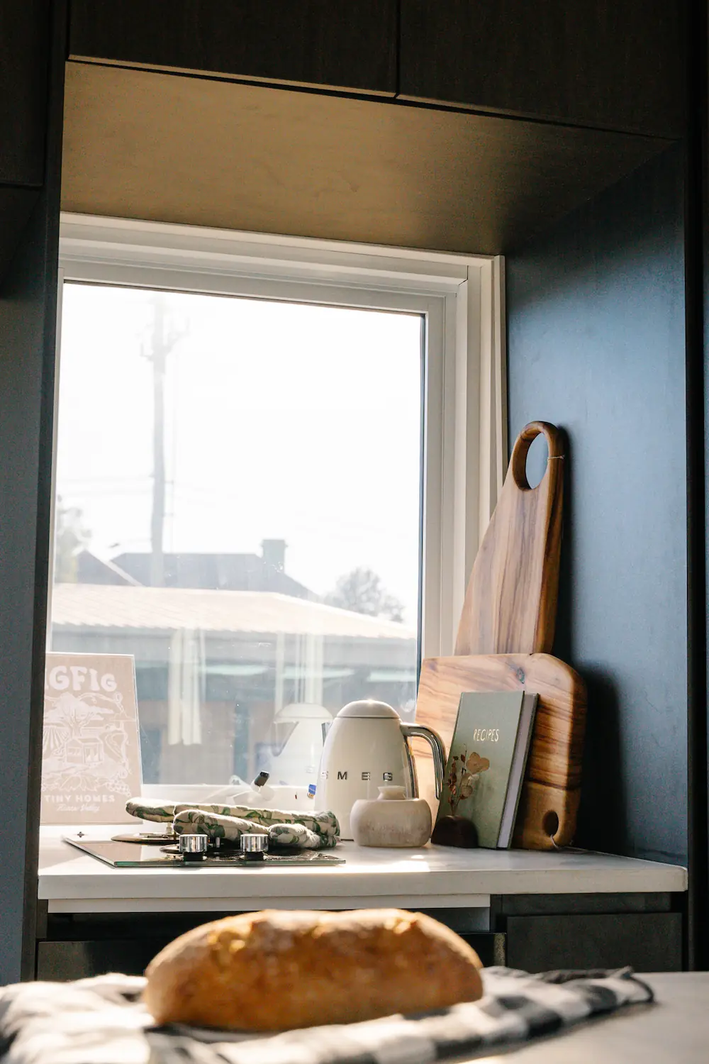 Kitchen window of Tiny Home countertop with a wooden cutting board, recipe book, SMEG kettle, and oven mitts, with a loaf of bread in the foreground.