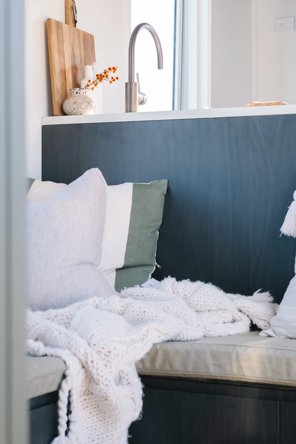 Cozy kitchen corner in a Tiny Home with a white knitted throw blanket and soft pillows on a cushioned bench under a countertop with a modern faucet and decorative items.
