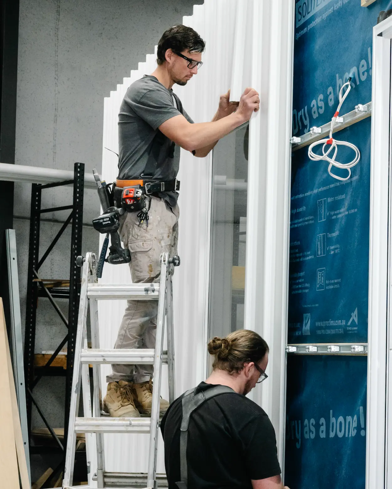 Two construction workers installing white siding panels on a building exterior, one on a ladder and the other preparing materials below.