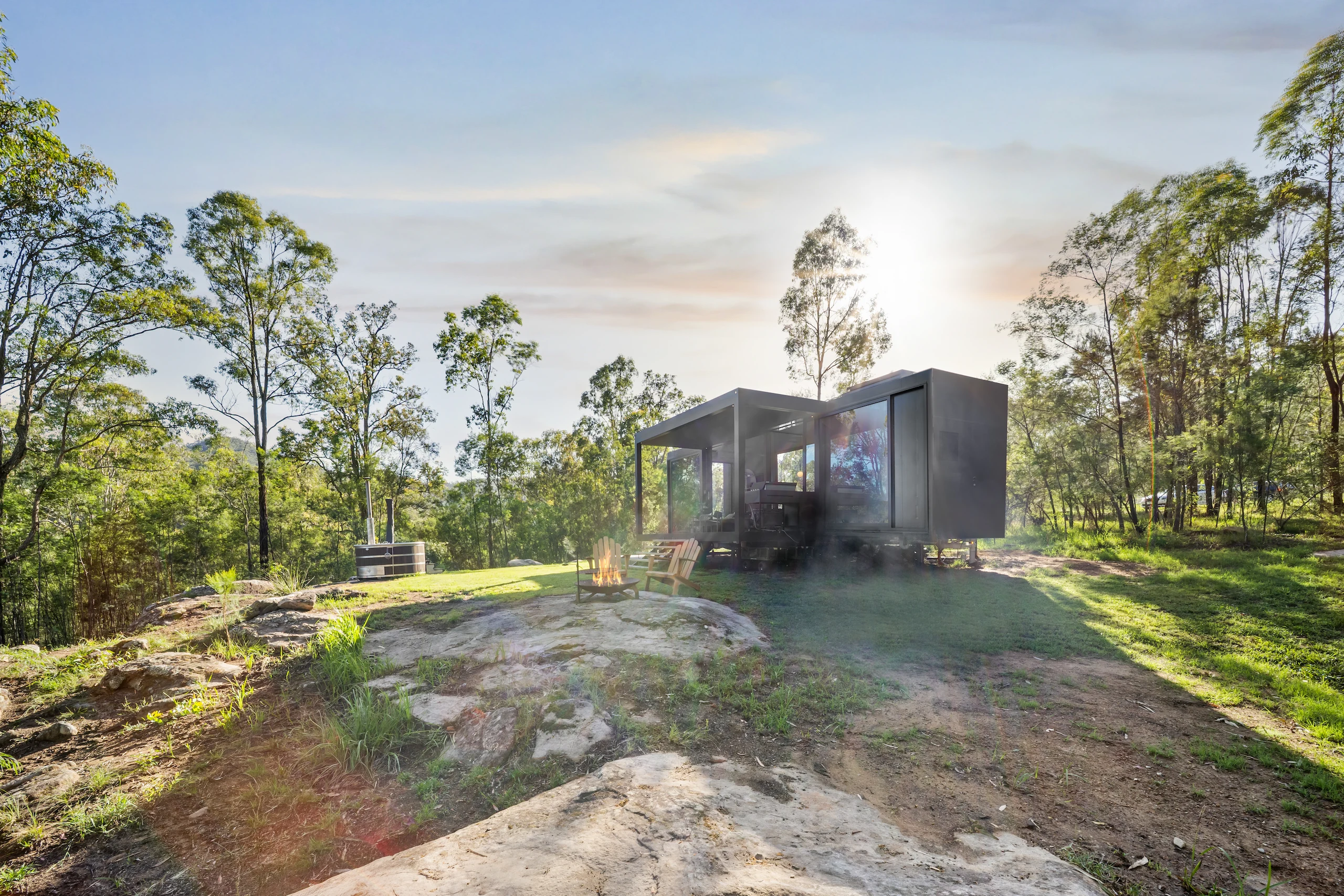 Modern Tiny Home with large windows surrounded by trees, featuring a fire pit with two wooden chairs on a rocky grassy clearing at sunset.