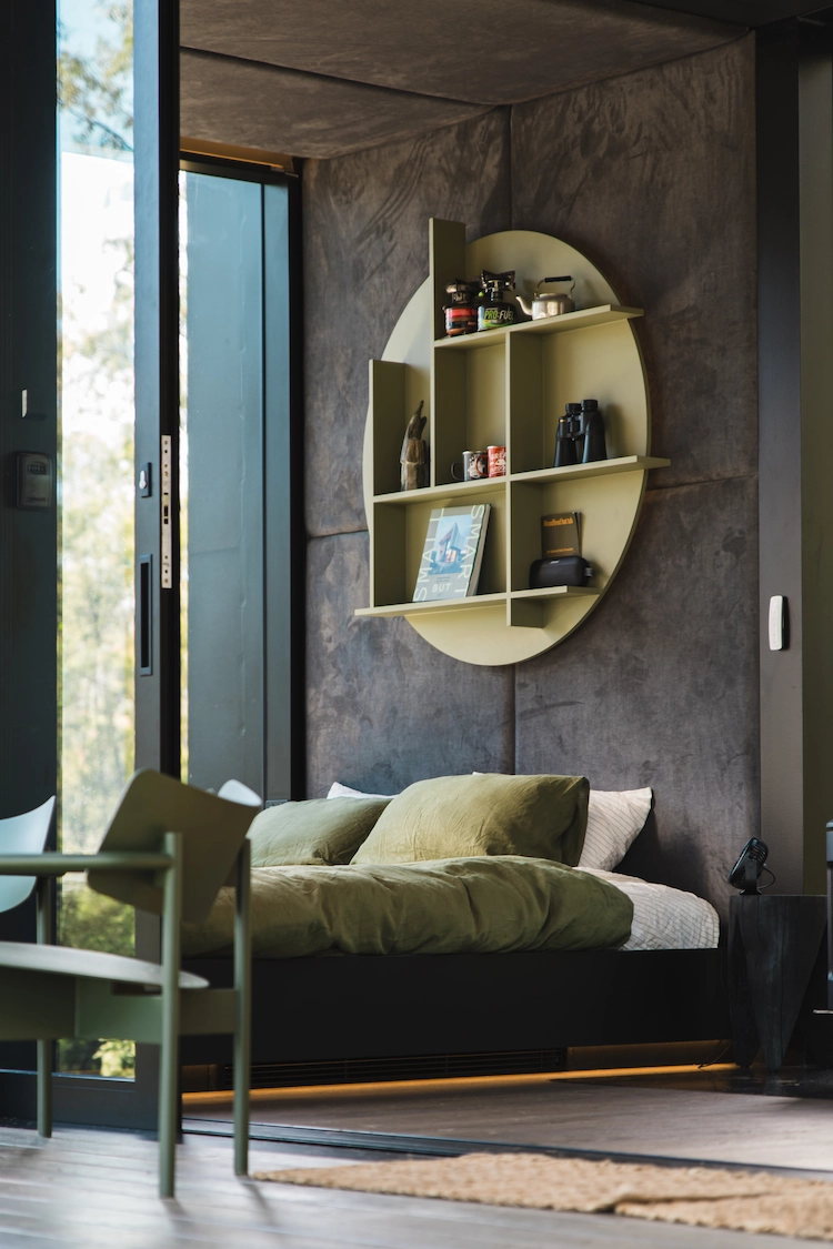 Modern bedroom in tiny home with dark walls, a green bed with matching pillows, a circular wall shelf above the bed holding books and decor, and a sliding glass door letting in natural light.