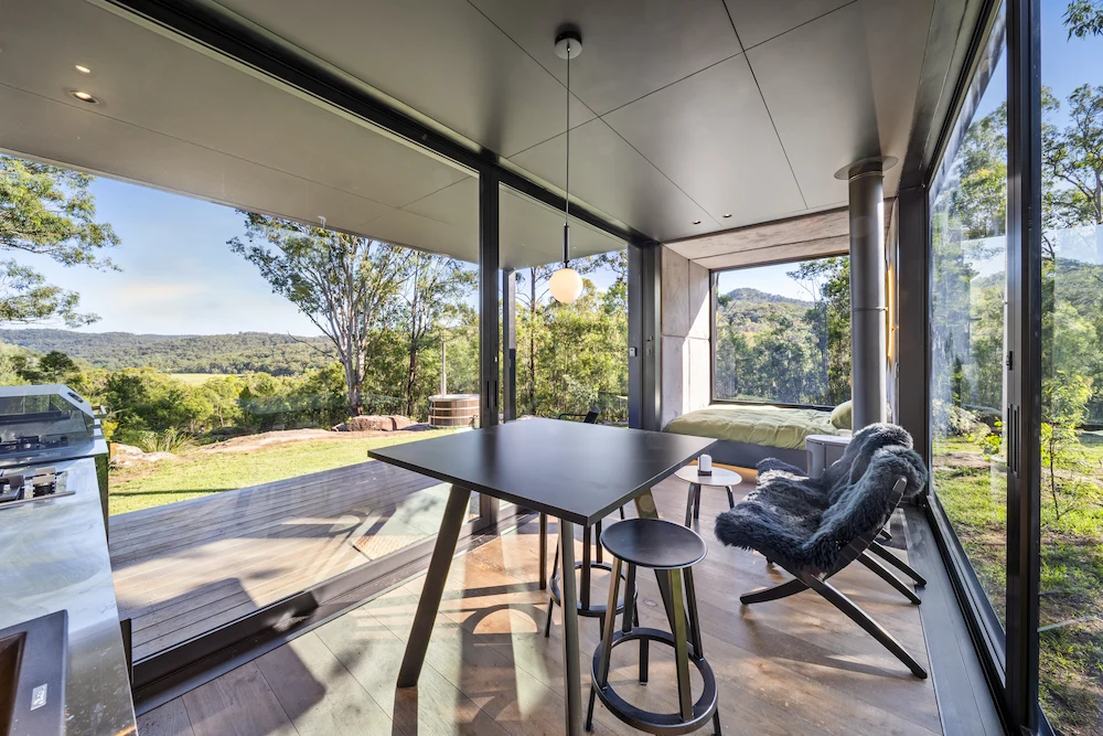 Modern glass-walled room with a table, stools, lounge chair, and a bed, overlooking a green forest and hills.