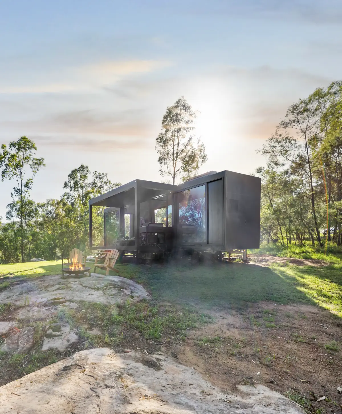 Modern black tiny home with large glass windows set in a forest clearing with two wooden chairs around a fire pit.