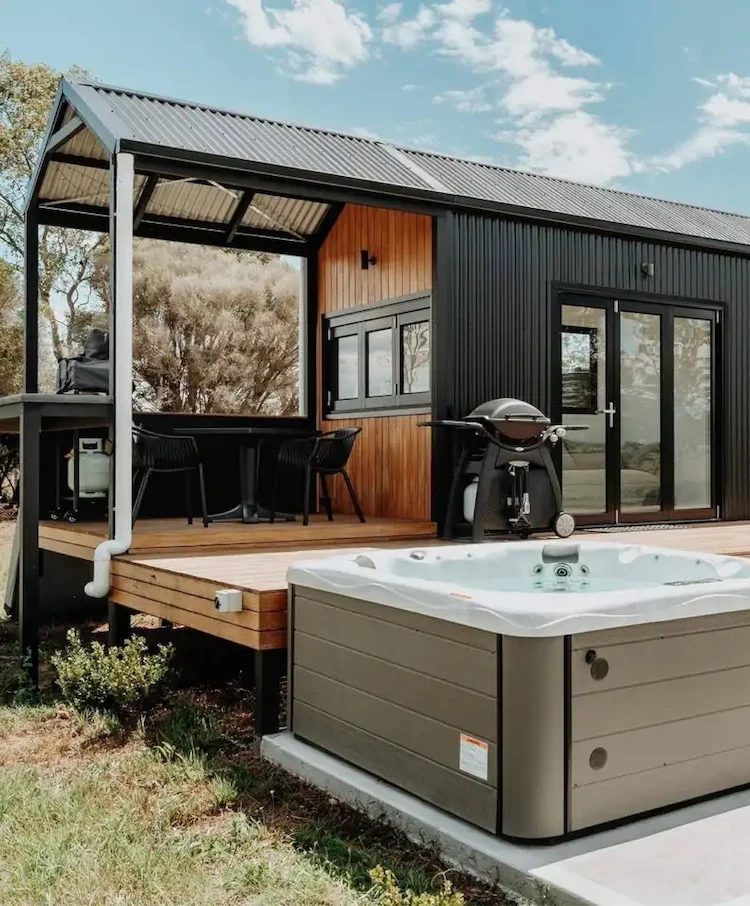 Modern small cabin with black exterior, wooden deck, outdoor seating, barbecue grill, and a hot tub in the foreground.