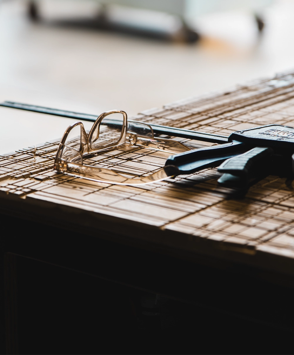Transparent eyeglasses and a black clamp resting on a wooden surface with a grid pattern.