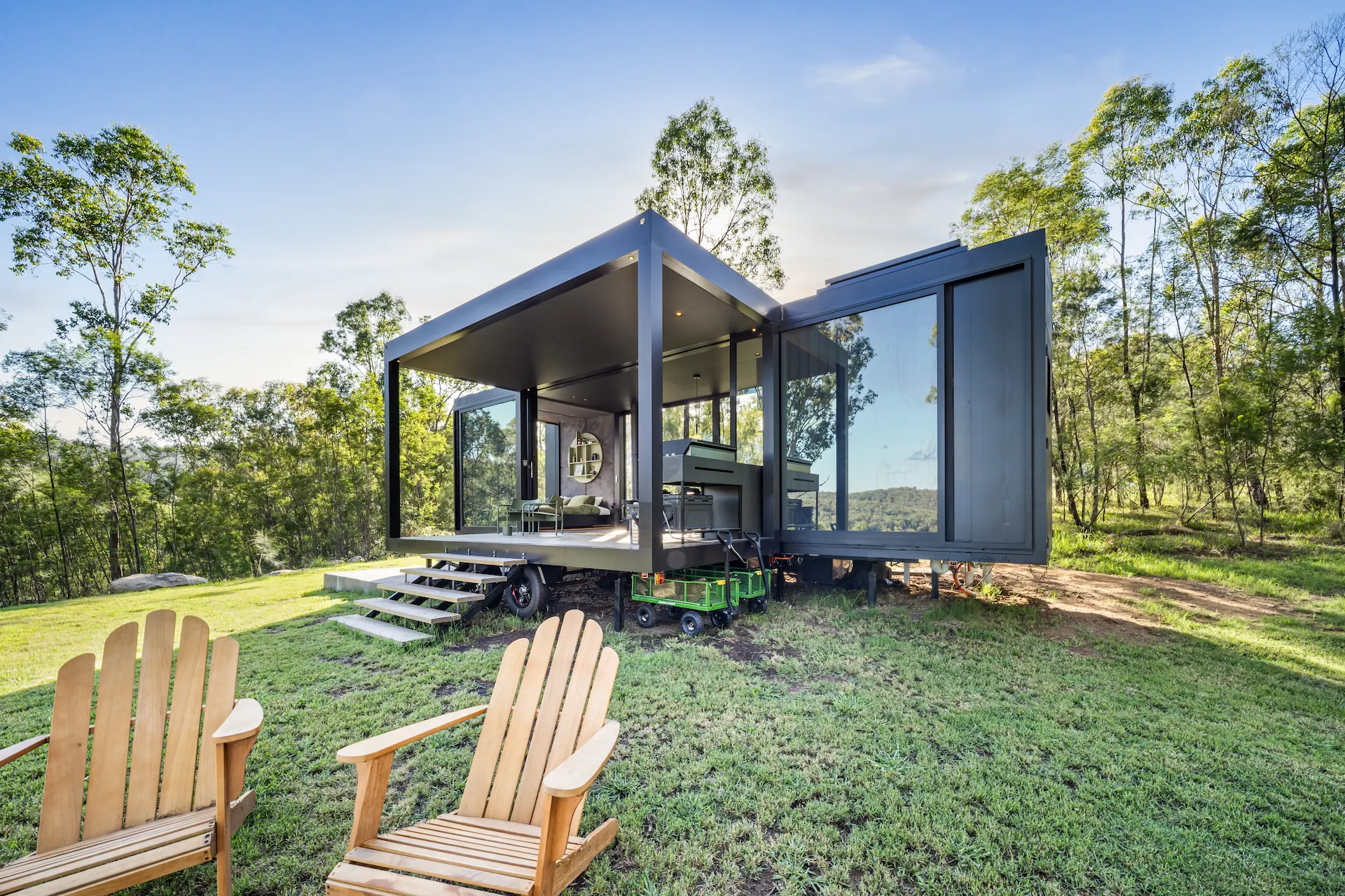 Modern black tiny house with glass walls and an open porch set in a grassy forest clearing with two wooden Adirondack chairs in the foreground.