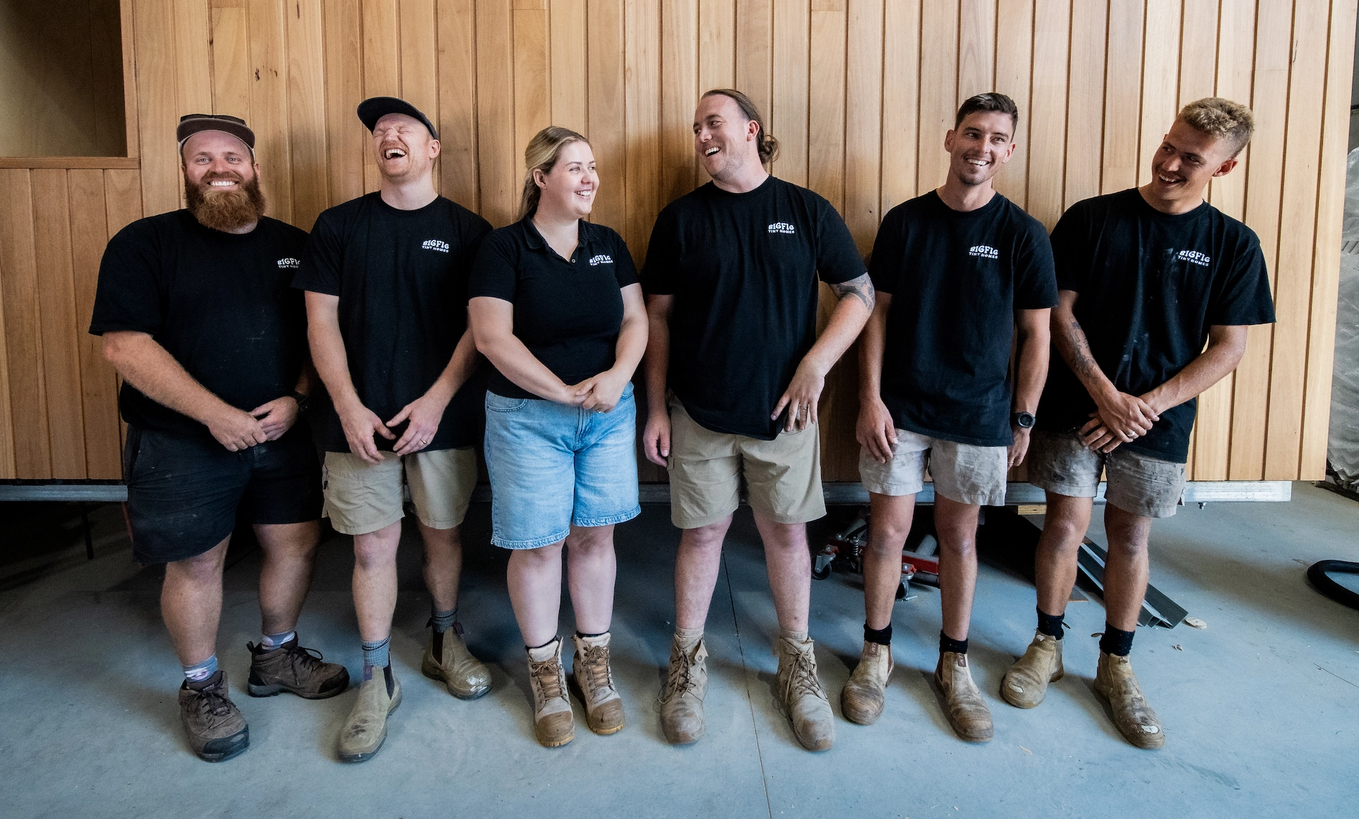 The Big Fig Tiny Homes team smiling workers wearing black company t-shirts and work boots standing against a wooden wall.