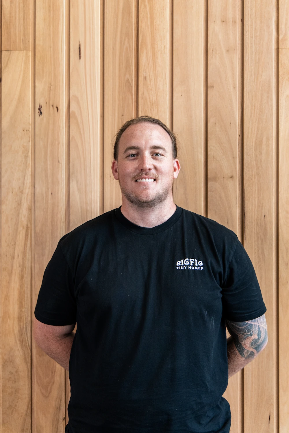 Smiling man with tattooed arm wearing a black Big Fig Tiny Homes t-shirt standing against a wooden panel wall.