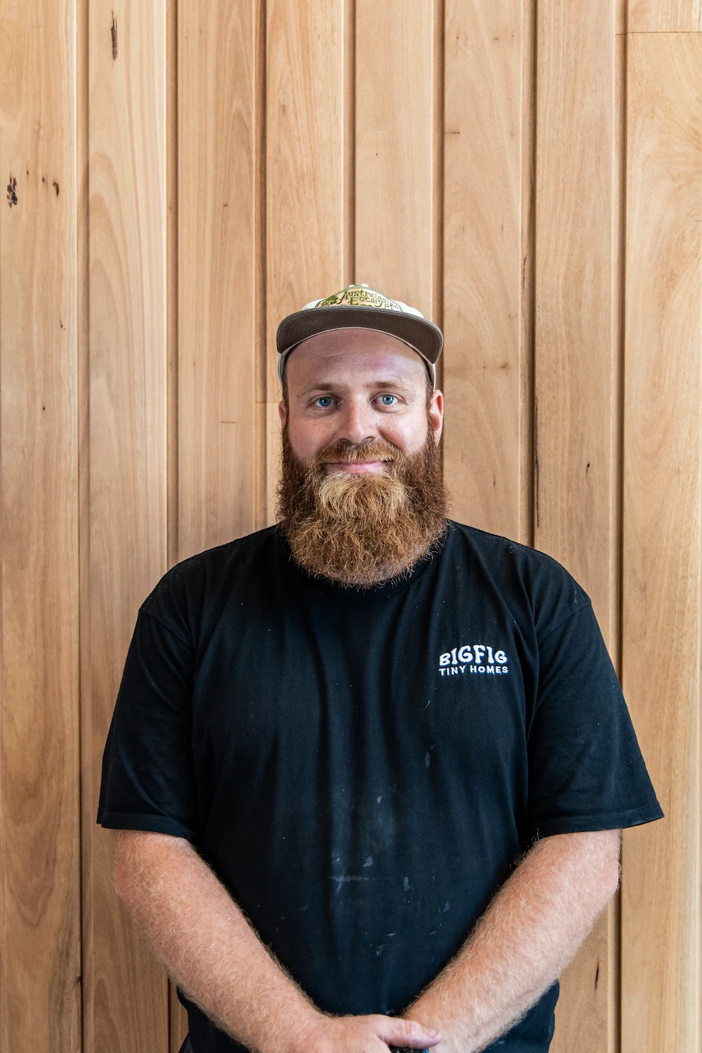 Bearded man wearing a cap and black BigFig Tiny Homes t-shirt standing in front of a wooden wall.