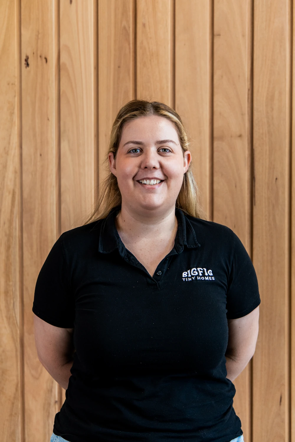 Smiling woman with blonde hair wearing a black BigFig Tiny Homes polo shirt standing in front of a wooden panel wall.