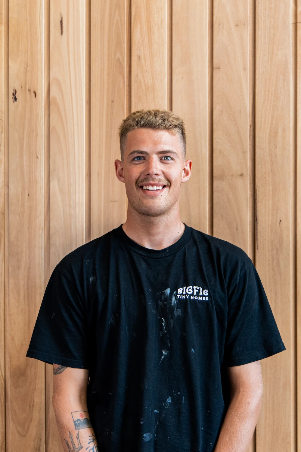 Smiling young man with short blond hair and a mustache wearing a black BigFig Tiny Homes T-shirt, standing against a wooden panel background.