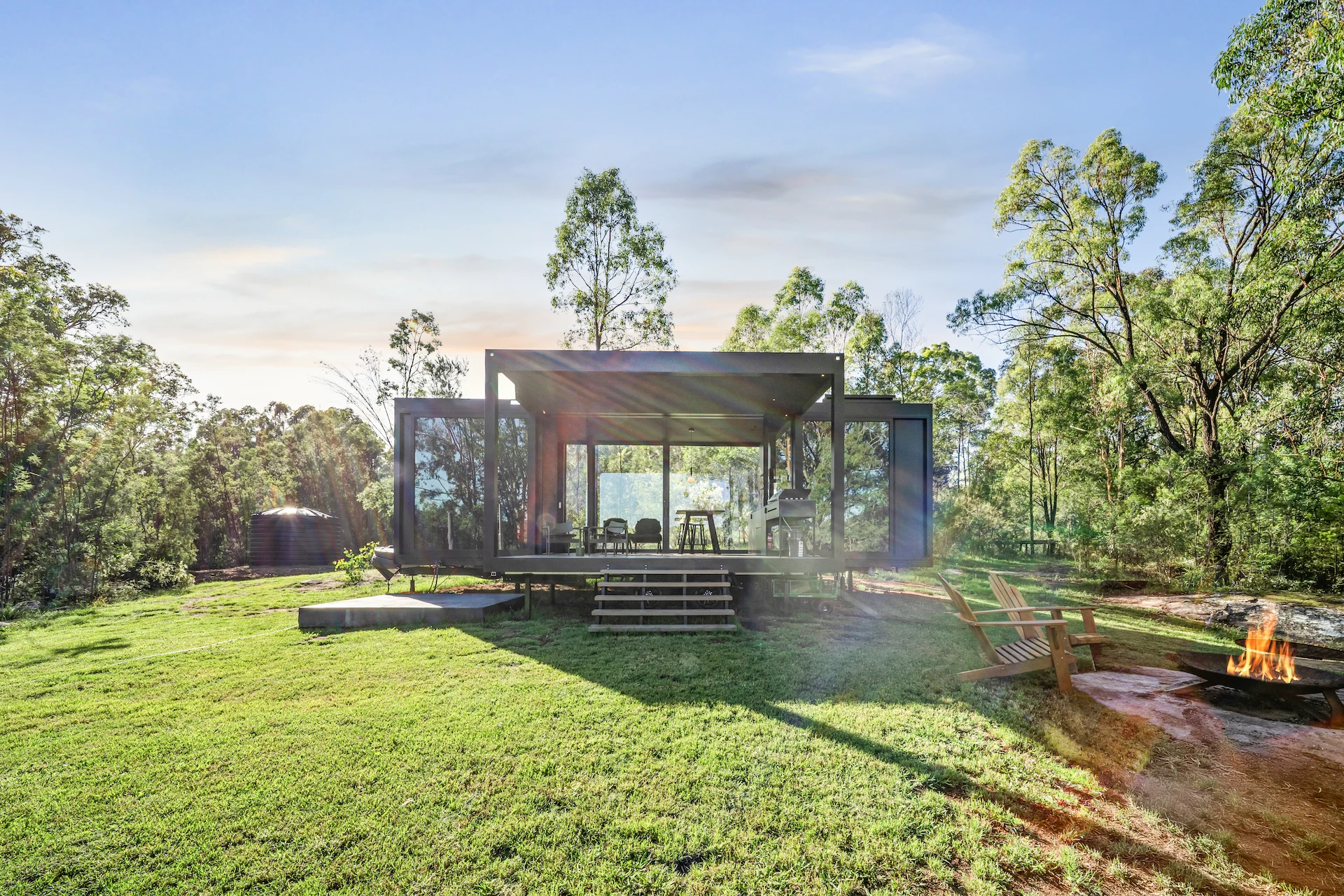 Modern glass Tiny Home on stilts in a grassy clearing surrounded by trees, with two wooden chairs and a fire pit nearby.