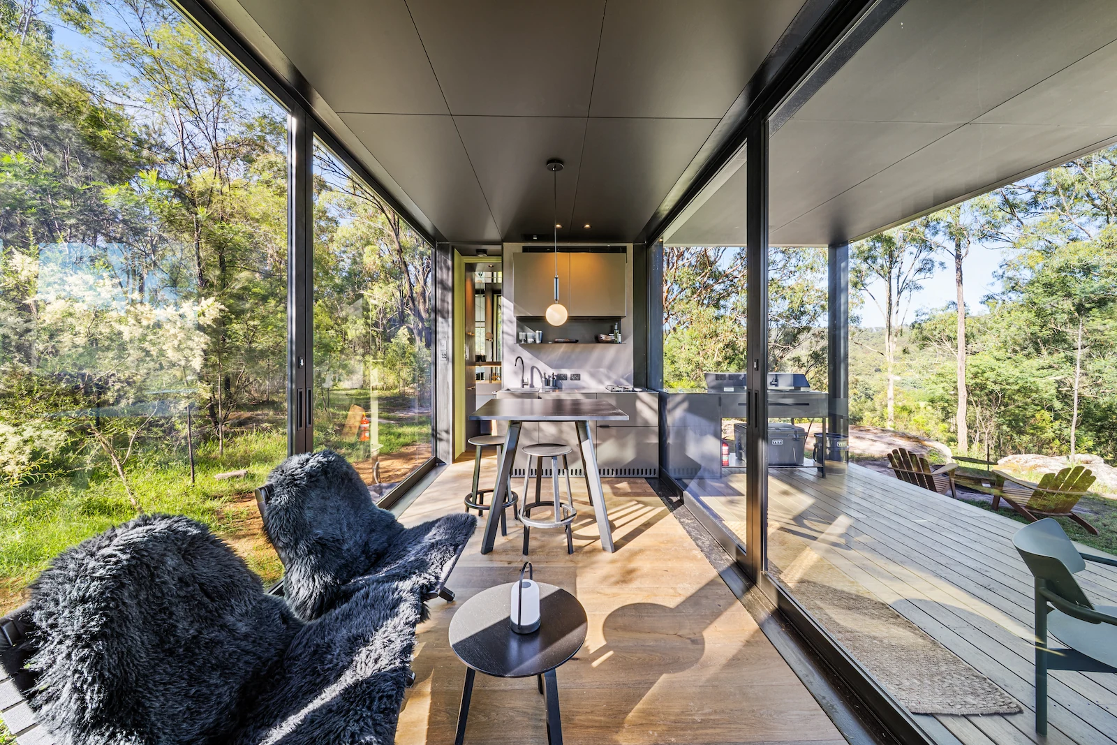 Modern small kitchen and seating area with large glass windows overlooking green forest and outdoor deck with chairs.