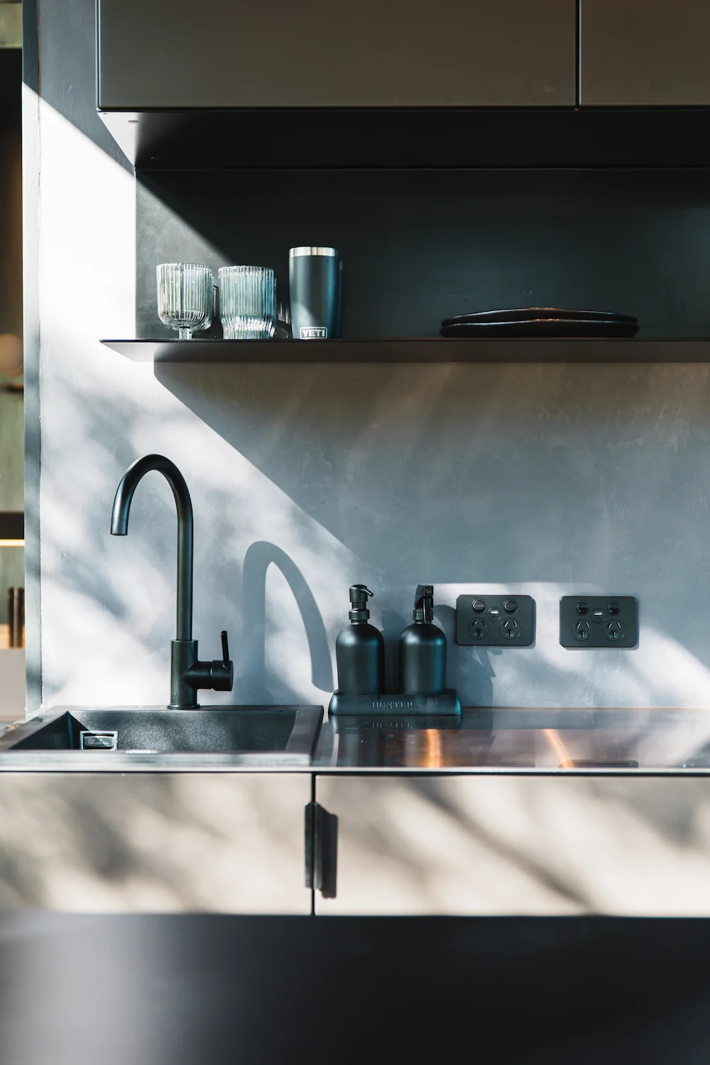 Minimalist kitchen sink area with black faucet, black soap dispensers, two power outlets, and shelf holding glassware and a YETI tumbler.