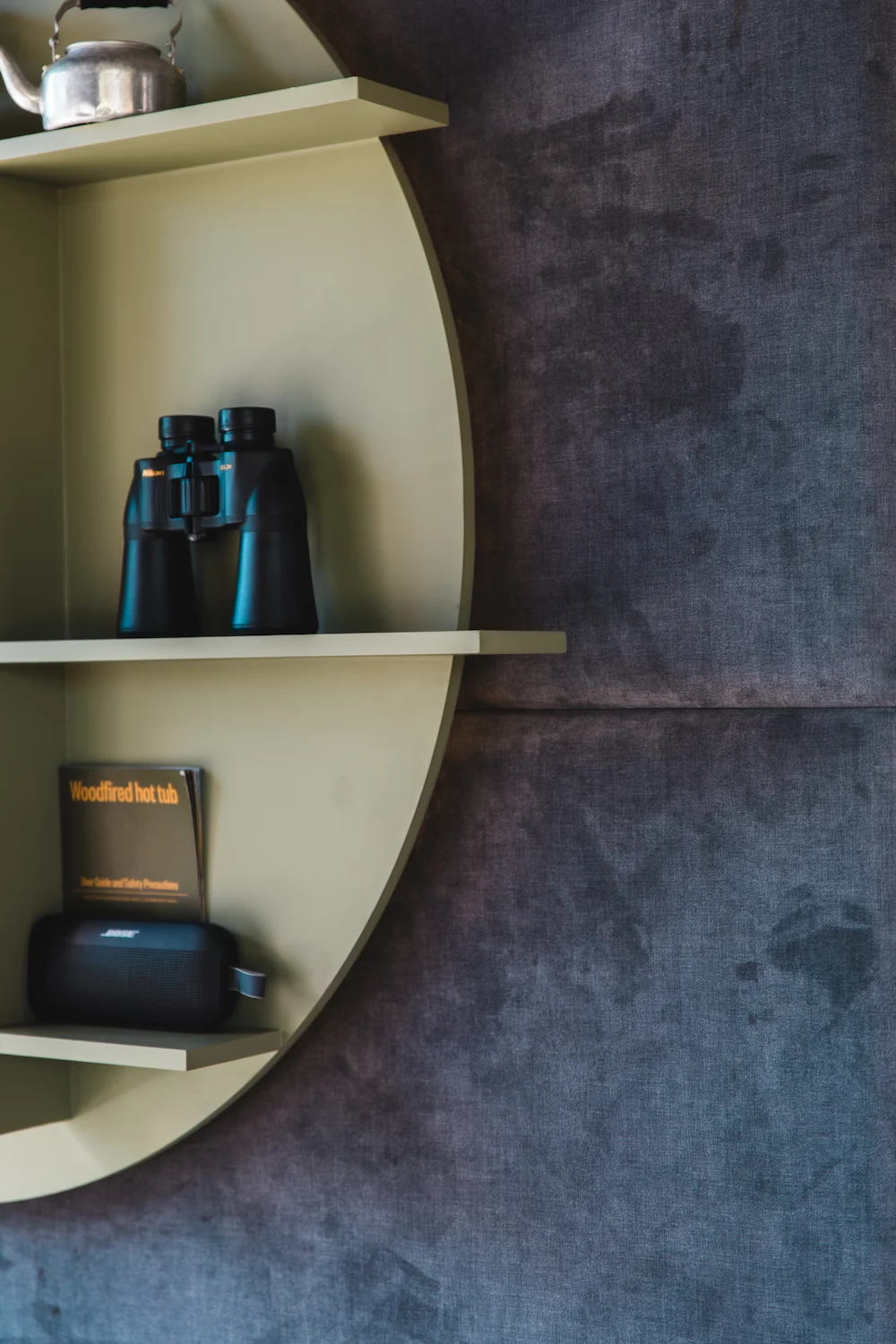 Circular beige shelf mounted on dark fabric wall with a silver kettle, black binoculars, a booklet titled 'Woodfired hot tub,' and a portable Bose speaker.