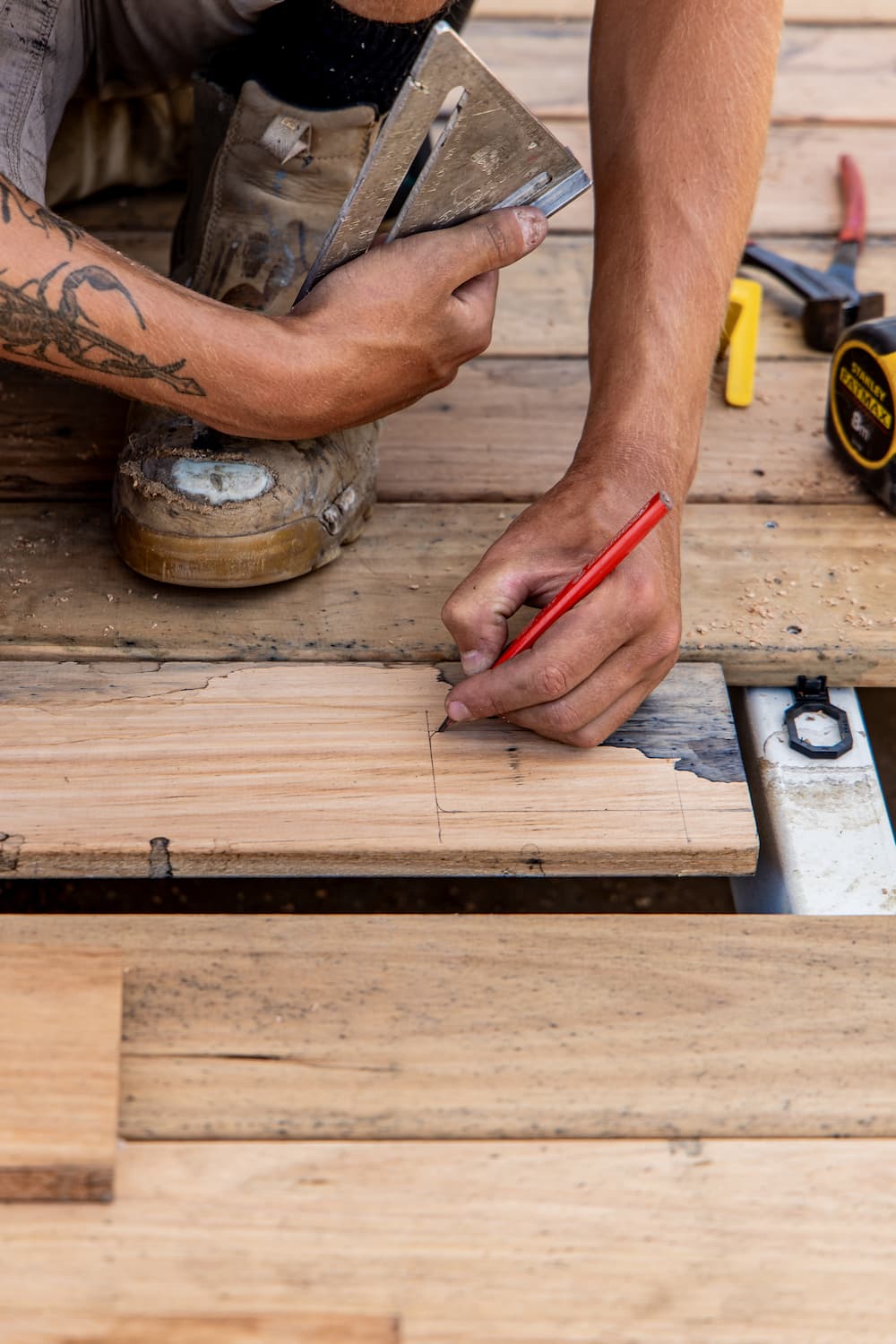Carpenter marking measurements on a wooden plank using a pencil and metal square on a construction site.
